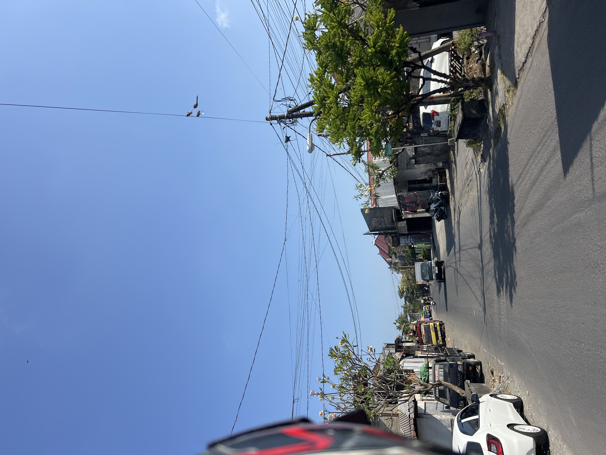 The image depicts a street scene in Denpasar, Indonesia, under a clear blue sky. The road is paved and appears to be a two-lane street with parked vehicles and some traffic. On the left side of the road, a white car is parked, followed by a black truck, and then a yellow truck. The buildings lining the street are modest in structure, some appearing to be shops or residences with corrugated metal roofs. A large tree with green foliage is prominent on the right side of the road, casting shadows. Numerous overhead power lines crisscross the sky, supported by utility poles. In the distance, more vehicles, including motorcycles, are visible on the road. A few birds are perched on the power lines in the upper right portion of the image. The overall impression is of a typical urban street in a tropical climate.