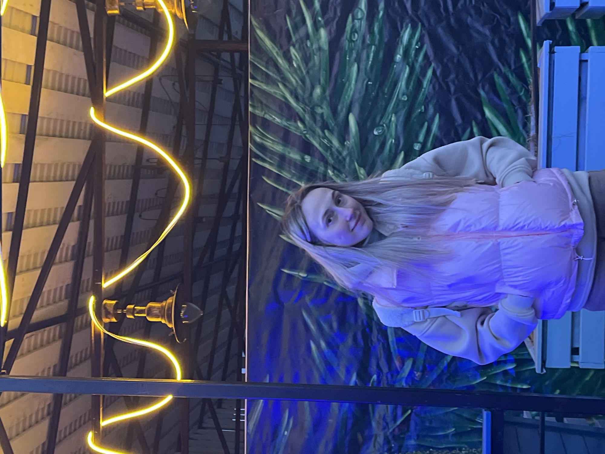 A woman with long blonde hair, wearing a light pink puffer jacket and a beige hoodie, is positioned in front of a background featuring green plant patterns with water droplets, and a blue light effect. A black metal railing is visible in the foreground, partially obscuring the lower part of the background. A decorative structure with a spiral of yellow lights and antique-style lighting fixtures is on the left. The photo appears to be taken in an urban setting.