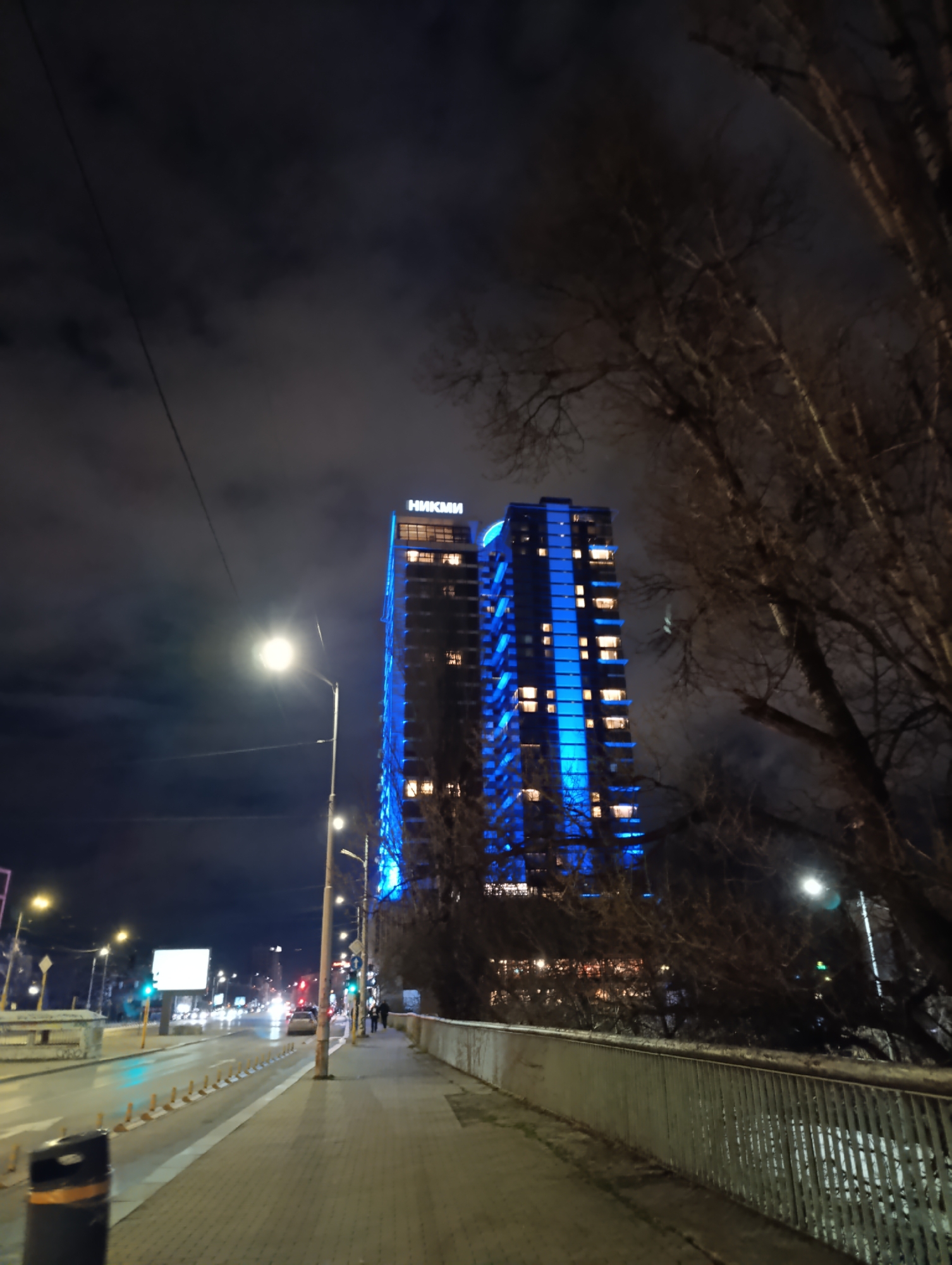 The image depicts a nighttime scene in Sofia, Bulgaria, featuring the Capital Fort business center, which is illuminated with blue lights. The view is from a sidewalk along a street lined with lampposts and traffic. Bare trees are visible on the right side of the frame. There are some pedestrians walking in the distance. A road runs parallel to the sidewalk, with cars and traffic lights visible. The sky is cloudy.