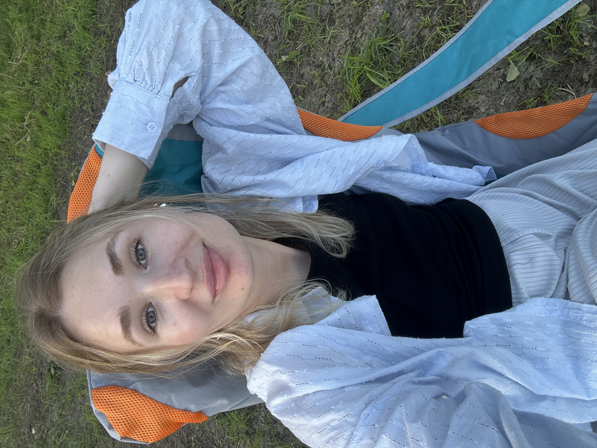 A young woman with blonde hair and blue eyes is sitting in a folding chair outdoors. She is wearing a black top and a light blue patterned shirt, with light-colored pants. Her right hand is resting on her head. The background appears to be a grassy area, consistent with an outdoor park or recreational space. The location is identified as Noviy, Russia.