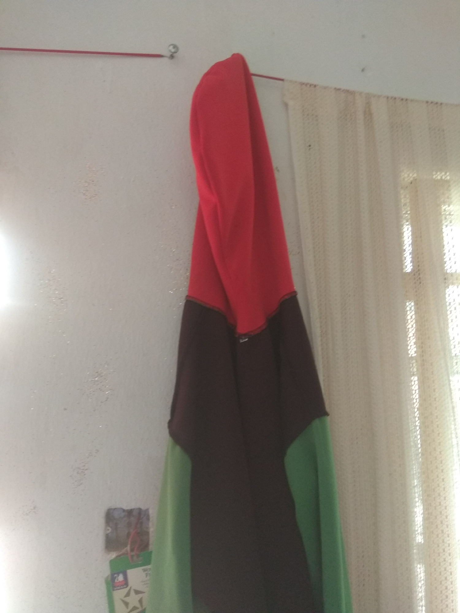 A close-up shot shows a red, black, and green garment hanging on a wall. The garment appears to be a hoodie, with the red portion forming the top, followed by a black middle section and green sleeves. The fabric is draped, creating folds and shadows that highlight its texture. To the left of the garment, a white wall is visible, with a red string and a small silver hook near the top. The wall has a textured surface with scattered specks that catch the light. Further to the left, a bright light source creates a luminous glow. On the wall, below the hanging garment, a visible electrical outlet with exposed wires and a green label attached to it. To the right of the garment, a cream-colored, sheer curtain with a textured pattern hangs, obscuring a window. The overall lighting suggests it might be daytime, given the bright light and the light filtering through the curtain. The scene appears to be indoors, likely in a domestic setting in Jalingo, Nigeria. No people are visible in the image. There is some visible text on the green label attached to the outlet, but it is not legible.