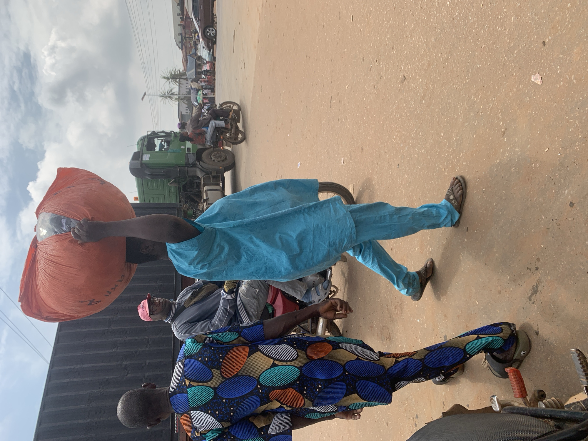 In Shagamu, Nigeria, an individual is seen walking on a dusty, paved road, carrying a large, orange, bagged object balanced on their head. The person is wearing a bright blue, loose-fitting garment. To the left, another person is walking away from the camera, wearing a vibrantly patterned, multi-colored top and pants. In the background, a green truck is parked, and a motorcycle with two passengers is in motion. The environment appears to be an urban street scene with buildings and trees visible further in the distance, under a partly cloudy sky.