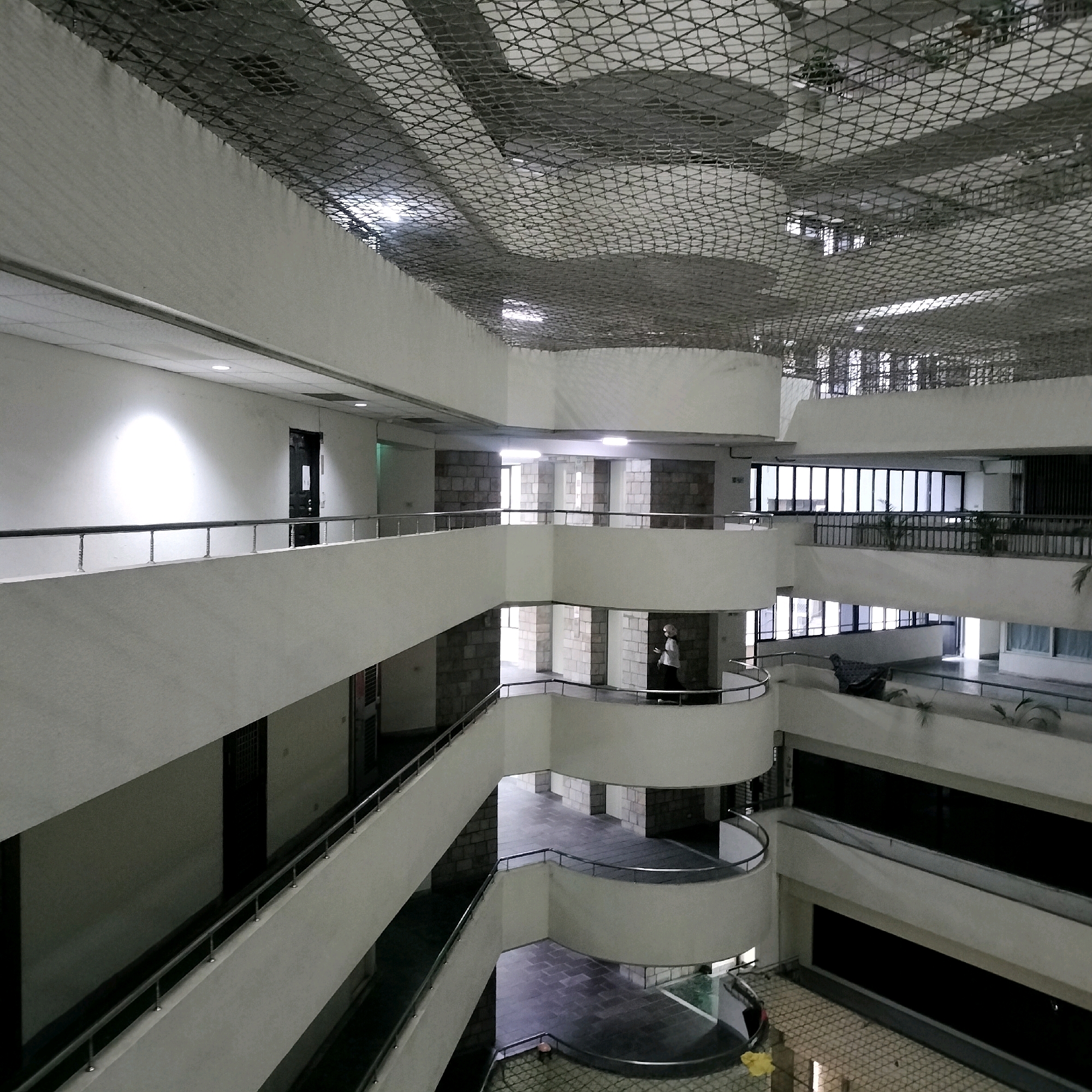 The image shows the interior of a multi-story building with a central atrium. The architecture features white, curved balconies and walkways, with metal railings. The ceiling is covered by a wire mesh, and lights are visible throughout. Several doorways and windows are present on the different levels. In the mid-ground, a person wearing a white head covering and dark clothing is walking on a curved balcony. The overall environment appears to be an indoor public space, possibly an office building, a convention center, or a museum, given the scale and design. Without further context or visible signage, pinpointing the exact location in Taichung, Taiwan, is not possible from the visual information alone.