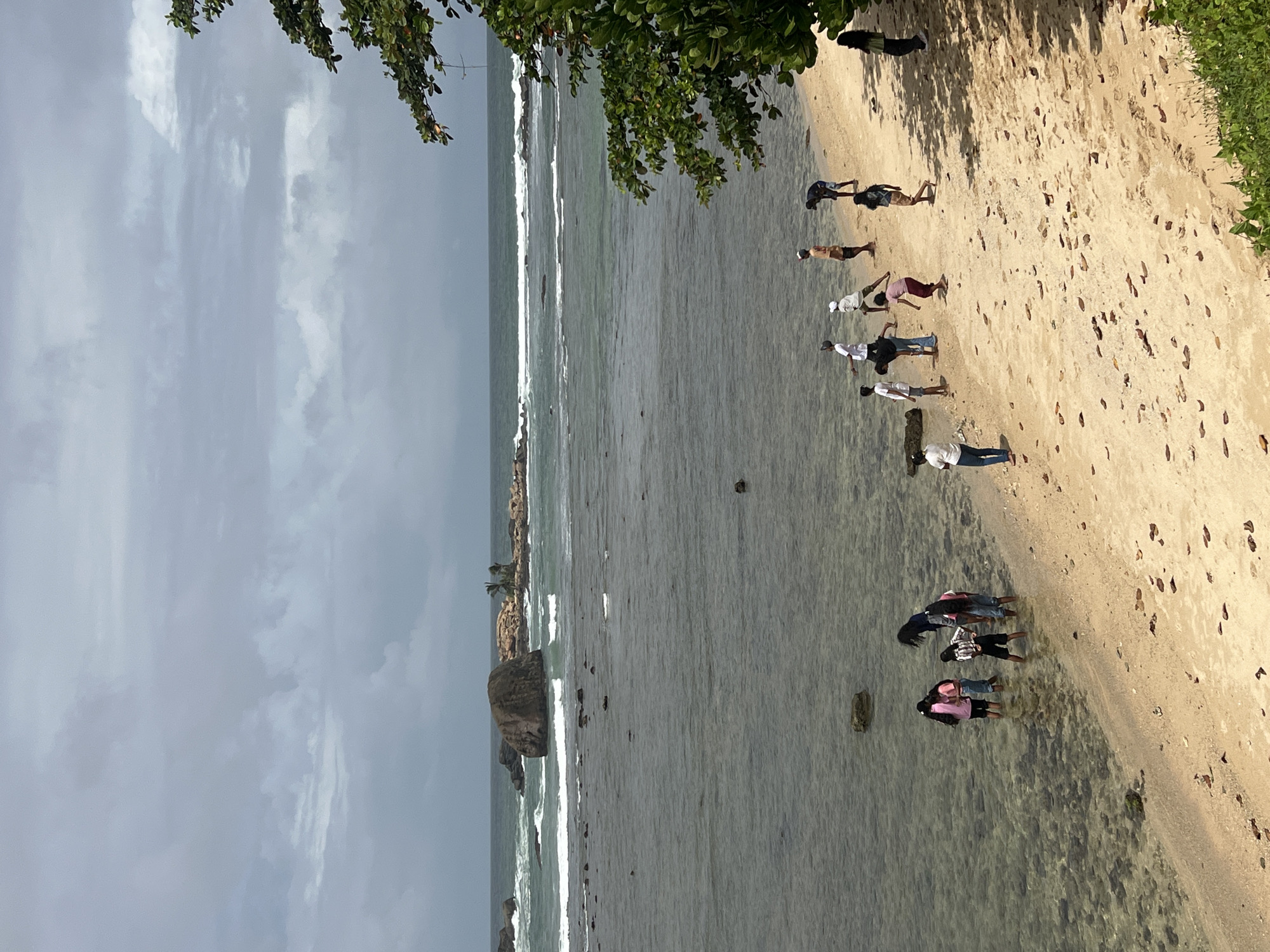 The image captures a wide shot of a rocky coastline in Galle, Sri Lanka, under a cloudy sky. In the foreground, a sandy beach is visible with scattered dry leaves. A group of people, appearing to be tourists or locals, are dispersed along the shoreline and in the shallow water. Some are standing on the beach, while others are wading in the clear, shallow water, exploring the rocky seabed. In the middle ground, the ocean is calm with gentle waves rolling towards the shore, revealing submerged rocks. Further out in the ocean, larger rock formations and a small island with palm trees are visible. The background is dominated by a grey, overcast sky. A lush green tree with dark leaves frames the right side of the image, casting shadows on the sand. The overall scene depicts a relaxed, outdoor activity at a coastal location.
