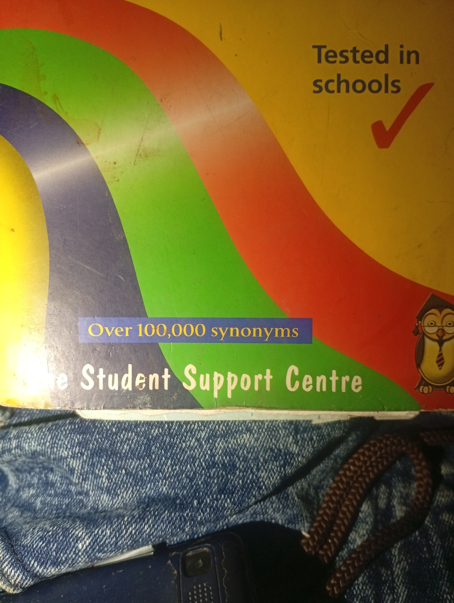The image shows a portion of a book cover with a colorful design featuring curved bands of yellow, orange, green, and blue. Text on the cover reads "Tested in schools" with a red checkmark, and "Over 100,000 synonyms" on a blue background. Below this, the text "e Student Support Centre" is visible in white lettering. A cartoon owl wearing a graduation cap and tie is present on the right side of the cover. The book cover appears to be placed on a dark blue denim fabric, possibly the leg of a pair of jeans, with a dark brown cord visible in the lower right section. The lighting suggests an indoor environment. There is no indication of a location in Gulak, Nigeria.