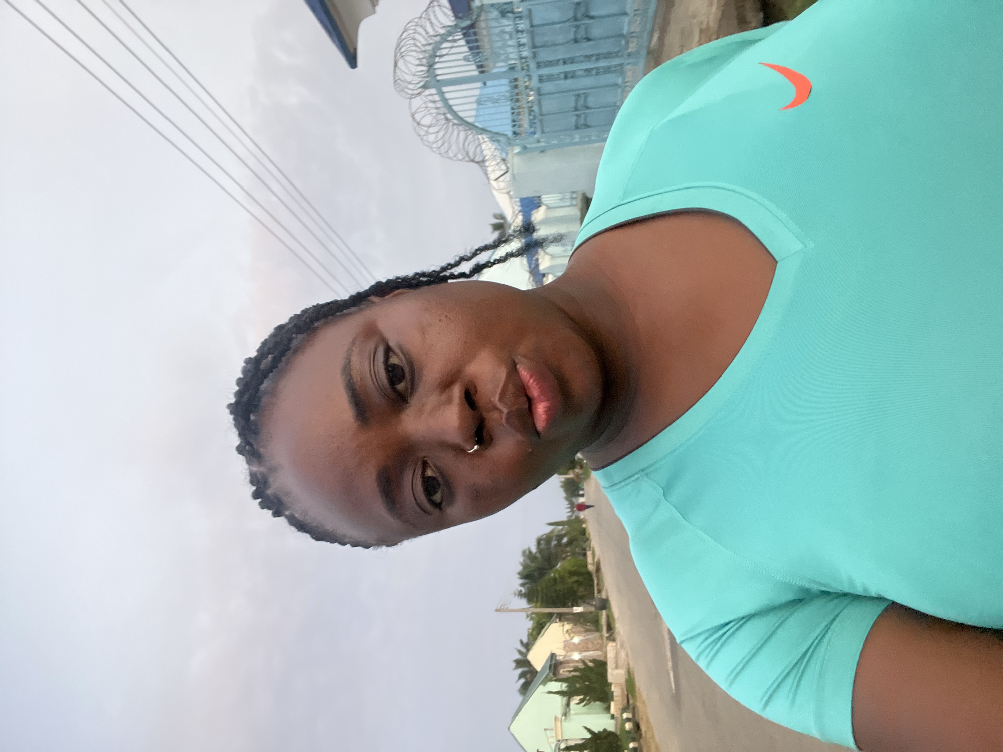 A medium-close-up selfie shows a Black woman with braided hair and a nose ring posing outdoors in what appears to be a residential area in Abuja, Nigeria. She is wearing a turquoise t-shirt with a Nike swoosh logo on the right side of her chest. Her lips are pursed as she looks at the camera. The background is slightly blurred but shows houses and a street, with power lines visible against a cloudy sky. A blue metal gate with barbed wire decorates the right side of the frame. The woman's expression is playful, and she seems to be taking the photo herself. The overall impression is that of a casual outdoor moment captured during a walk.