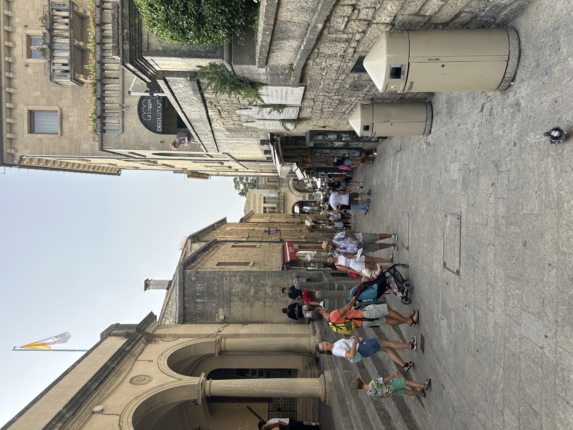 A group of tourists are walking along a cobblestone street in Città di San Marino, San Marino. The street is lined with stone buildings, some of which have arched doorways and balconies. A flag, likely the flag of San Marino or the Vatican, flies from a pole on one of the buildings. People are dressed in casual summer clothing. Several are pushing strollers, and a young boy is walking ahead of his family. There are shops with awnings lining the street, and on the right side of the image, there are two large beige trash receptacles. The sky is clear and bright.