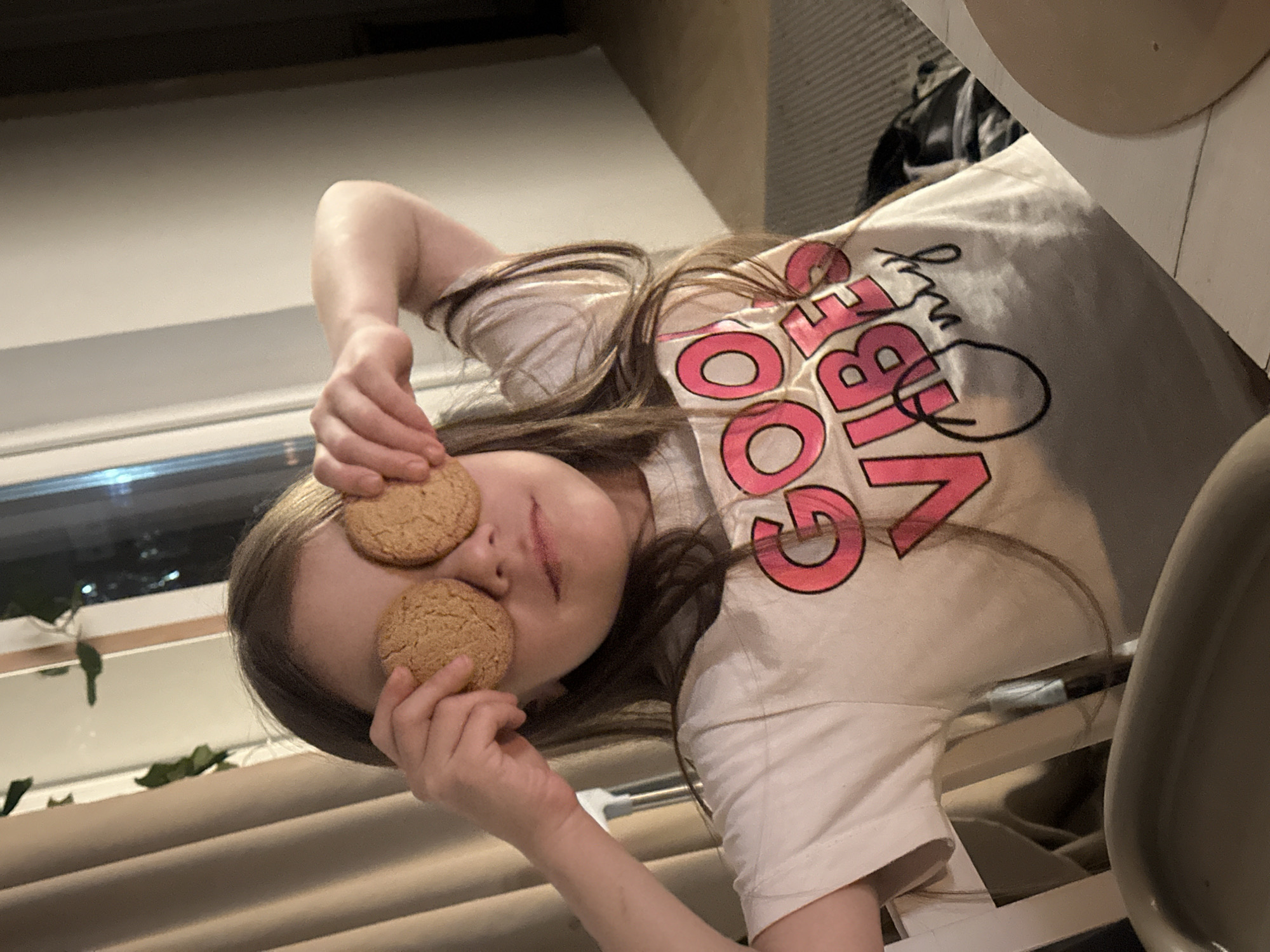 A young girl is holding two round cookies over her eyes in an indoor setting. The girl has brown hair and is wearing a light-colored t-shirt with pink and black lettering that says "GOOD VIBES". She is holding the cookies with her hands, one cookie over each eye. The background includes window blinds, a window with some greenery visible outside, and part of what appears to be a chair and possibly a countertop. The location is not discernable based solely on the image.
