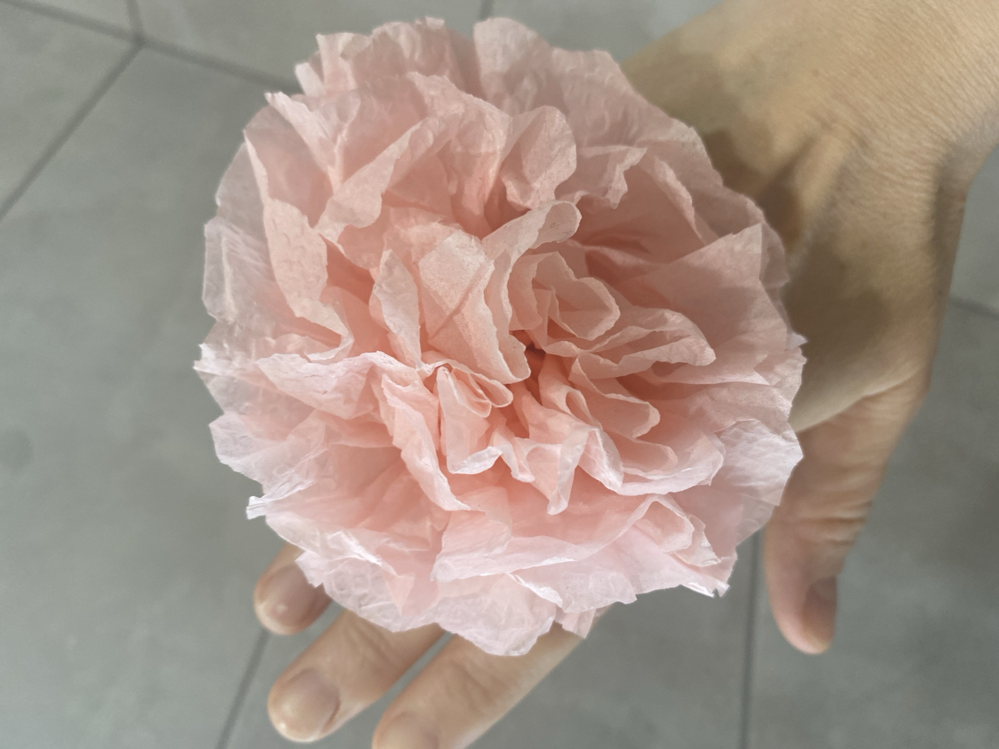 A hand holds a pink paper flower against a light gray tiled background. The hand is light-skinned and partially obscured by the flower. The flower is large and composed of multiple layers of delicate, crumpled paper petals. No further information about the location is discernible from the image.