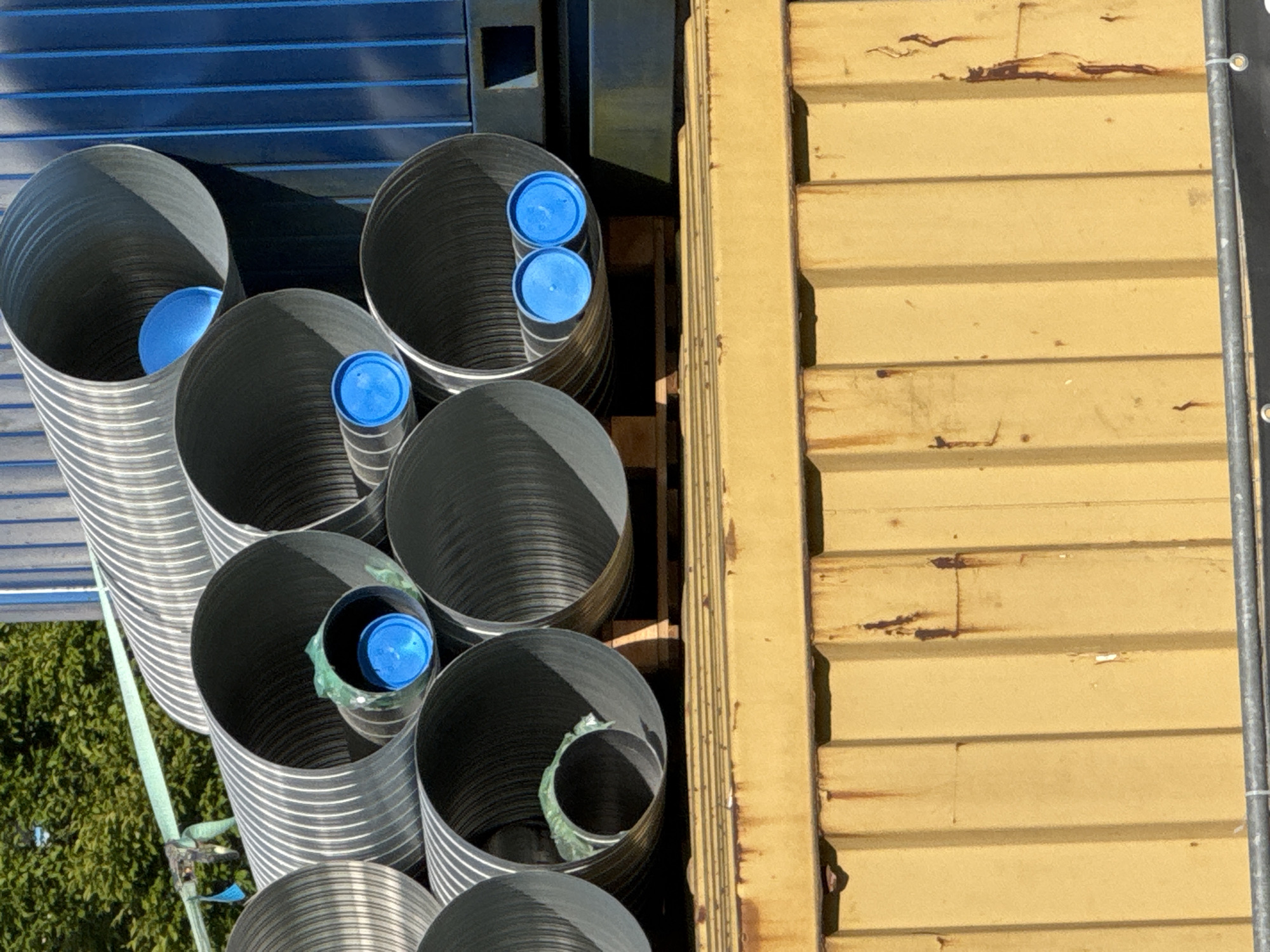 The image shows a close-up, overhead view of several large, corrugated pipes stacked together. The pipes are made of a silver-colored material and have a ribbed texture. Some of the pipes are capped with bright blue plastic lids, while others are open. Inside one of the open pipes, a piece of green plastic material is visible.
The pipes are arranged in a dense grouping, with their openings facing upwards. They appear to be stored outdoors, possibly at a construction site or a warehouse. To the right of the pipes is a section of a large, yellow corrugated metal container or structure, showing signs of rust and weathering. Behind the pipes, on the left side of the image, is a dark blue corrugated surface, likely another part of the storage area. A hint of green foliage from trees or bushes can be seen in the lower left corner, suggesting an outdoor setting with some vegetation nearby. A light blue strap is visible in the lower left, securing some of the pipes.
The lighting suggests it is daytime, with shadows cast by the pipes, indicating sunlight. The overall scene is functional and industrial, with no people visible. The focus is on the arrangement and details of the pipes and their immediate surroundings. The specific location is Höhenkirchen-Siegertsbrunn, Germany.