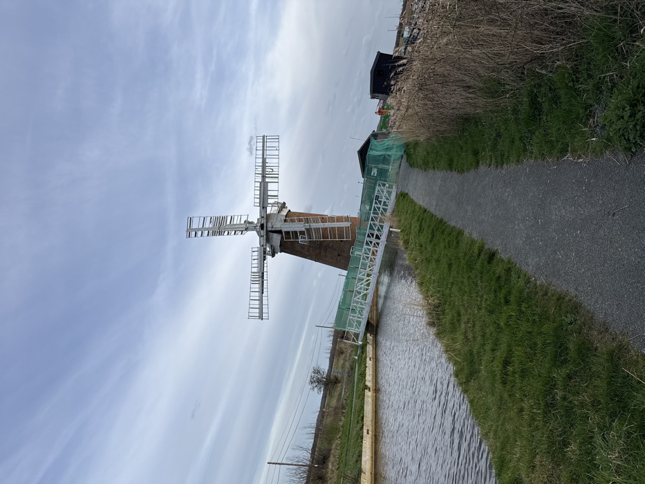 The image depicts Horsey Windpump in North Norfolk, United Kingdom, on an overcast day. A prominent brick-built windpump with four skeletal sails, devoid of fabric, stands in the midground, its base partially obscured by a temporary white metal gangway or bridge extending from the right bank across a dark body of water (likely a dyke or canal). This gangway is lined with light green netting. To the immediate right of the windpump, several temporary structures, including a dark-colored shed and other construction materials, are visible. In the foreground, a dark grey gravel path curves from the bottom right towards the center, bordered by green grass on its left and tall, dry reeds or grass on its right. The water body runs parallel to the path on the left, reflecting the muted sky. The background reveals a flat, sparsely vegetated landscape with distant trees and utility poles under a light grey, cloudy sky. No individuals are present in the scene.