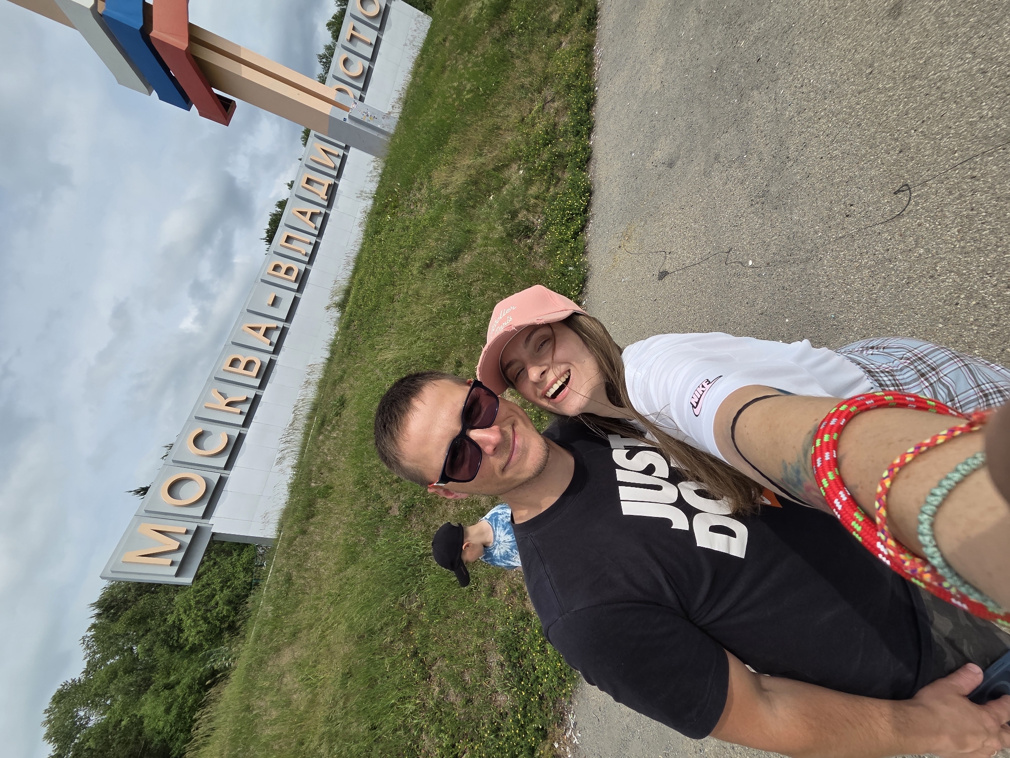In Nikolaevka, Russia, a young couple takes a selfie in front of a large sign that reads "MOSKVA-VLADIVOSTOK". The man, wearing sunglasses and a black t-shirt with "JUST DO IT" printed on it, smiles at the camera. The woman next to him, wearing a pink cap and a white t-shirt, laughs. A young boy in a black cap and a tie-dye t-shirt is visible in the background, walking up a grassy hill. The sign is positioned on the side of a road, with a vibrant green hill and trees behind it. A colorful, abstract sculpture stands to the right of the sign, with layers of red, blue, and white. The sky is cloudy, suggesting an overcast day.