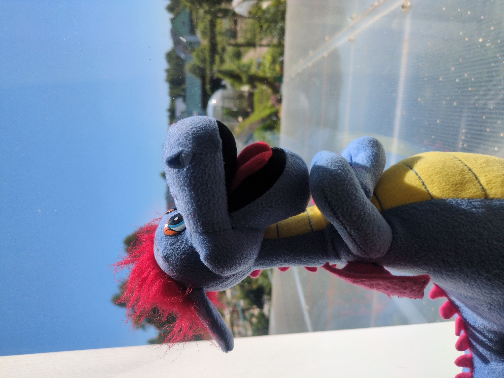 The image features a close-up, tilted shot of a blue stuffed dragon toy. The dragon has bright red, spiky hair, a yellow and blue striped back, and large blue paws. Its mouth is open, revealing a red tongue and black lining. The background is a blurred, sunlit scene with a clear blue sky, green trees, and a translucent, ribbed structure that appears to be made of plastic or glass. The lighting suggests it is daytime, possibly mid-morning or afternoon, with bright sunlight. The overall impression is a whimsical and possibly playful scene, perhaps set outdoors or near a window, given the light and background elements. There is no visible text in the image.