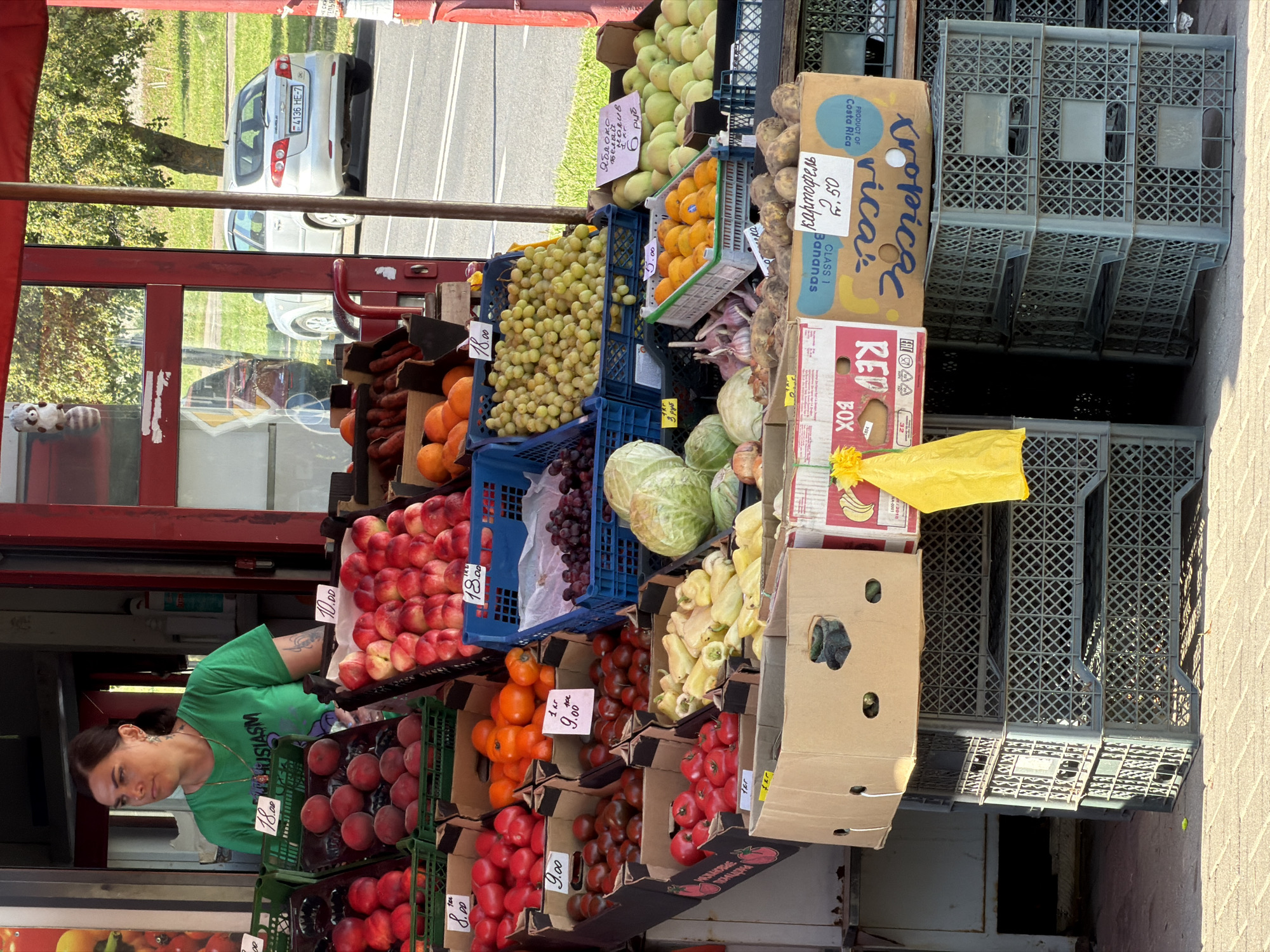 An outdoor fruit and vegetable stand is pictured with a woman standing behind it. The woman is wearing a green t-shirt and a necklace and has dark hair pulled back. She appears to be looking to her left. In front of her, an assortment of fruits and vegetables are displayed in crates and cardboard boxes. These include peaches, tomatoes, oranges, grapes, cabbage, peppers, garlic, and potatoes. Some of the produce has price tags. In the background, a silver car is parked on a street, and trees and grass are visible. The scene suggests a marketplace or roadside stall. The license plate of the car, "4136 HE-7," and the Cyrillic text on some of the signs and price tags indicate the location is likely Minsk, Belarus.