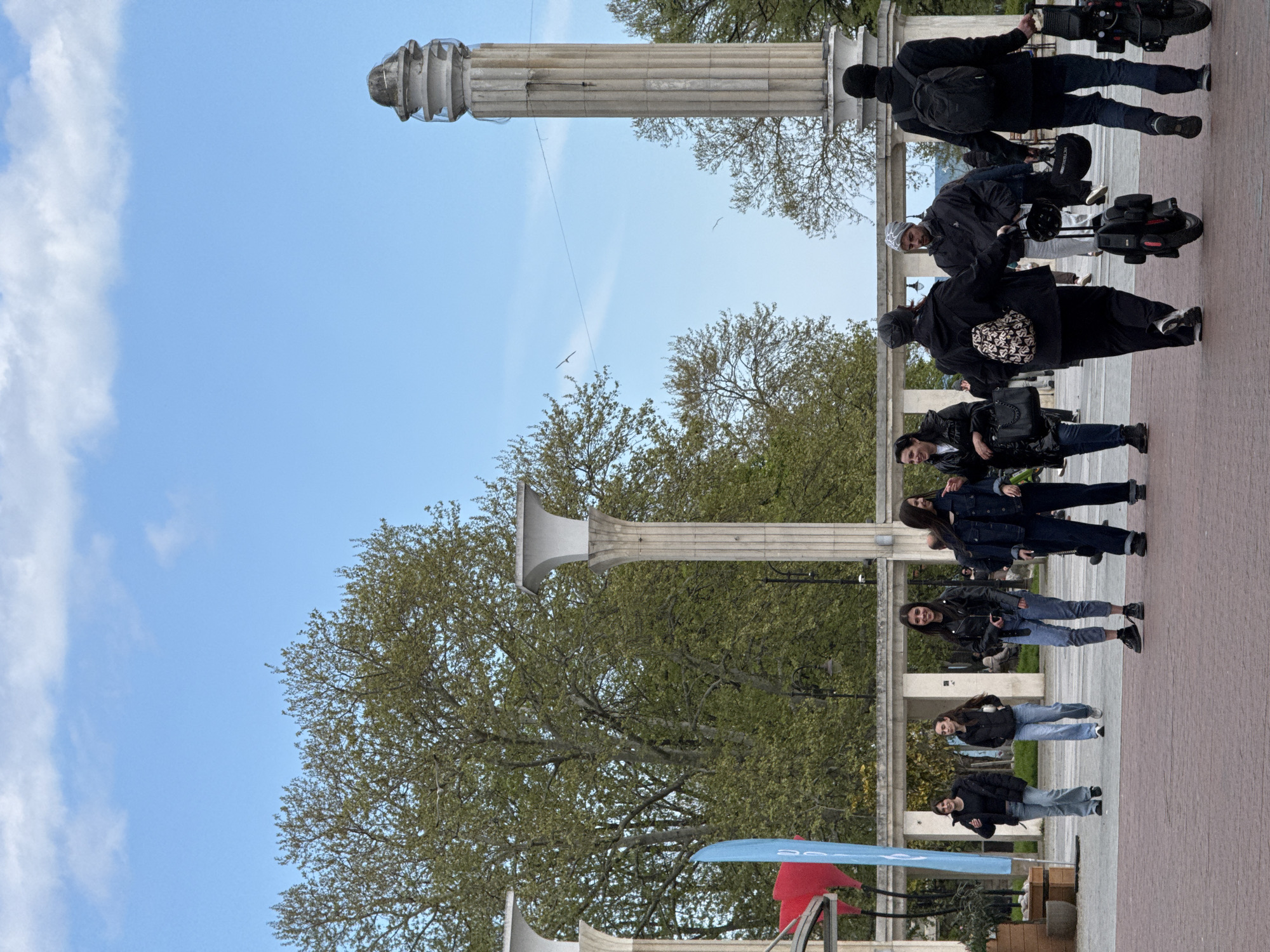 The image depicts a group of people on a paved square with columns. The location is Varna, Bulgaria, judging from the architecture and general appearance. A group of people riding electric unicycles are in the upper right corner. Several people are standing and posing for a photo, or simply observing the scene. A row of trees are visible in the background behind the columns. The sky is blue with some white clouds.