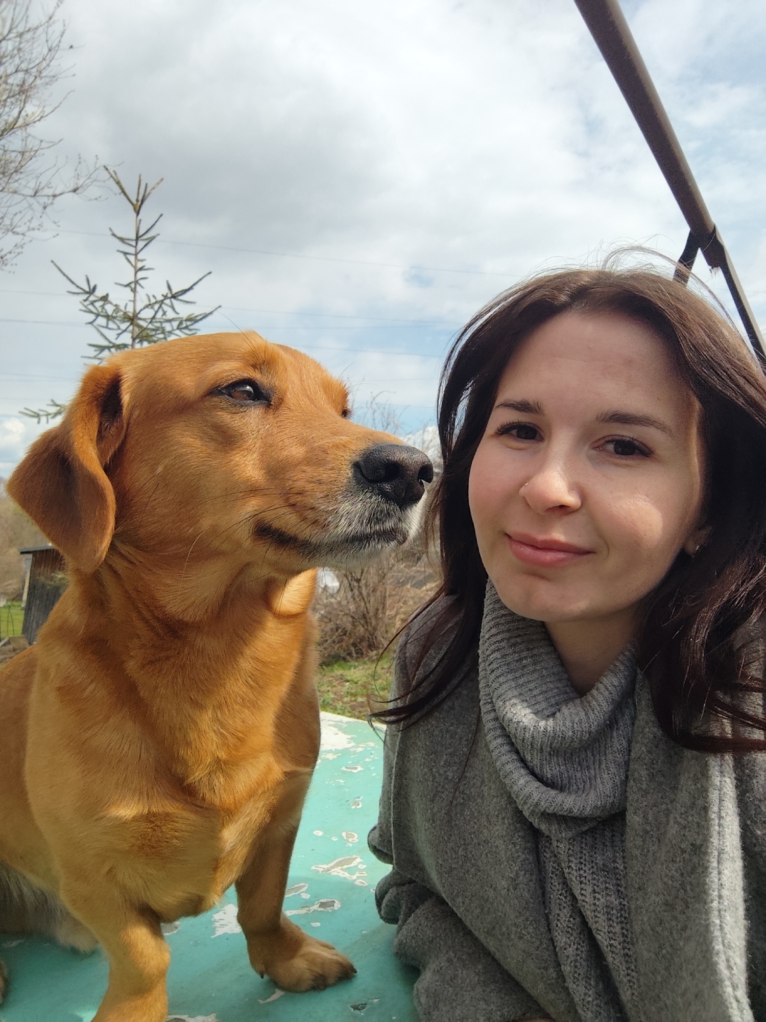 A woman with dark brown hair and a gray scarf poses next to a medium-sized, light brown dog with floppy ears. The dog is sitting on a turquoise surface. Behind them, a partly cloudy sky is visible, along with trees and a section of metal structure. The setting appears to be outdoors in an informal area, likely a yard or garden. The location is Фоки, Russia.