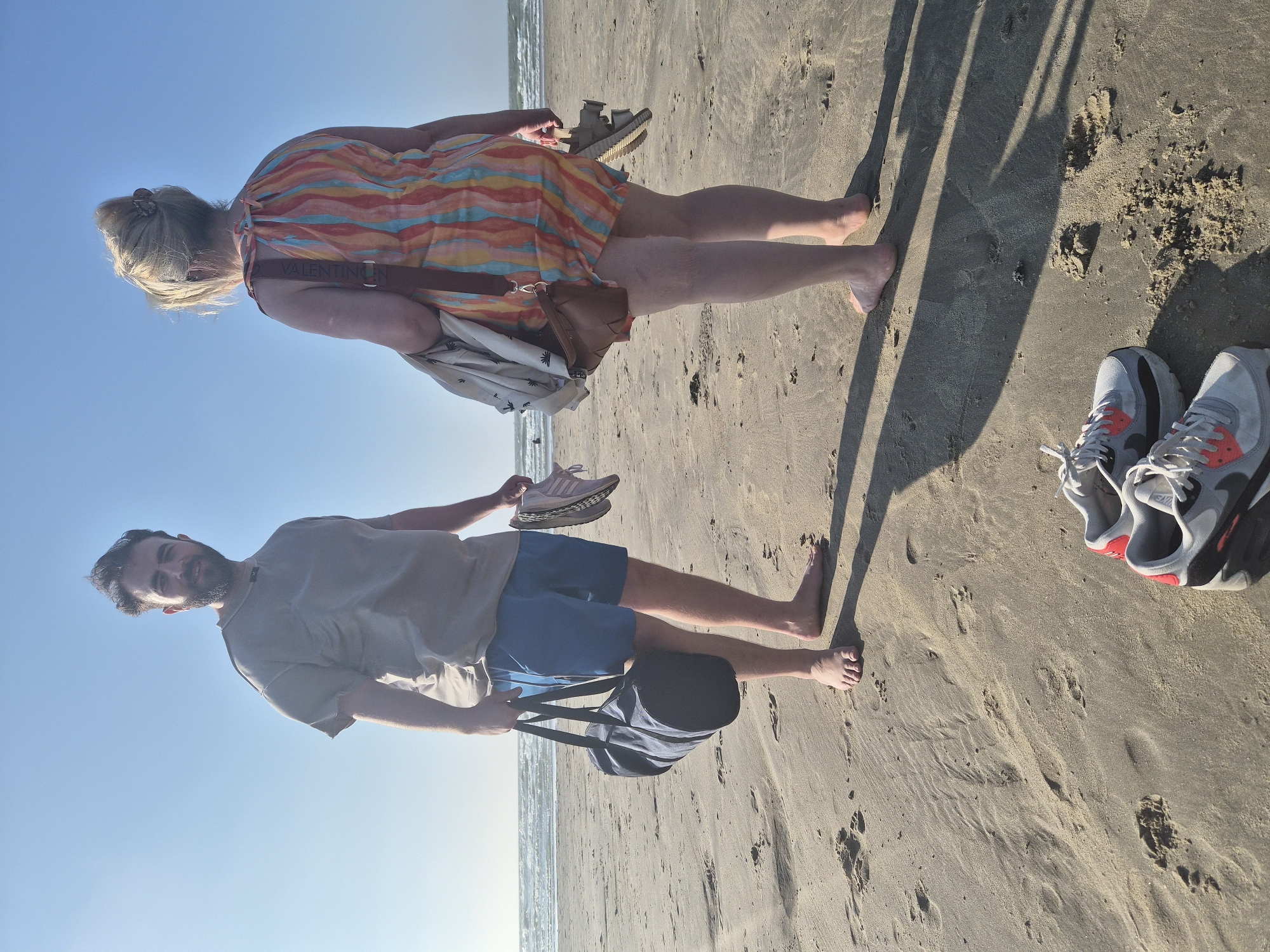 Two individuals are standing on a sandy beach with the ocean and a clear blue sky in the background. The man, on the left, is wearing a grey t-shirt and blue shorts. He is holding a dark blue duffel bag in his left hand and a pair of pink and white sneakers in his right hand. The woman, on the right, is facing away from the camera and is wearing a colorful striped dress. She is carrying a white jacket over her left arm and a brown purse slung across her body. In her right hand, she is holding a pair of beige sandals. In the foreground, a pair of grey and red Nike Air Max sneakers are placed on the sand. The image appears to be taken in the city of Blankenberge, Belgium, based on the provided context. The scene depicts a casual beach setting.