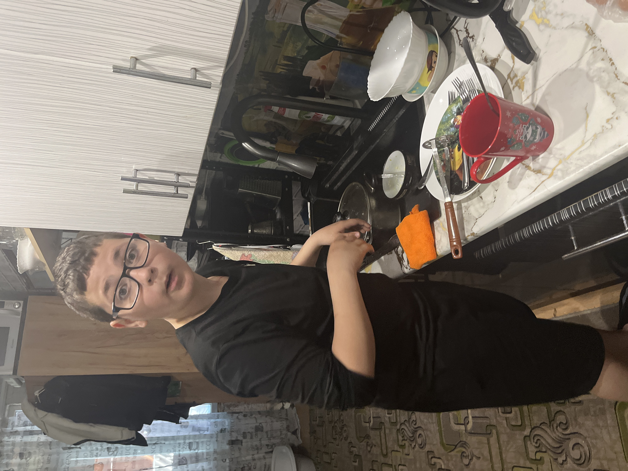 A person with short brown hair and glasses, wearing a black t-shirt, stands in a kitchen. The kitchen has white upper cabinets with horizontal bar handles. Above the sink is a dish rack holding dishes including a red mug, plates, bowls, and silverware. A black faucet is mounted to the sink. A small orange towel is visible. The floor has a patterned rug. The location is Vesyoloye, Russia.