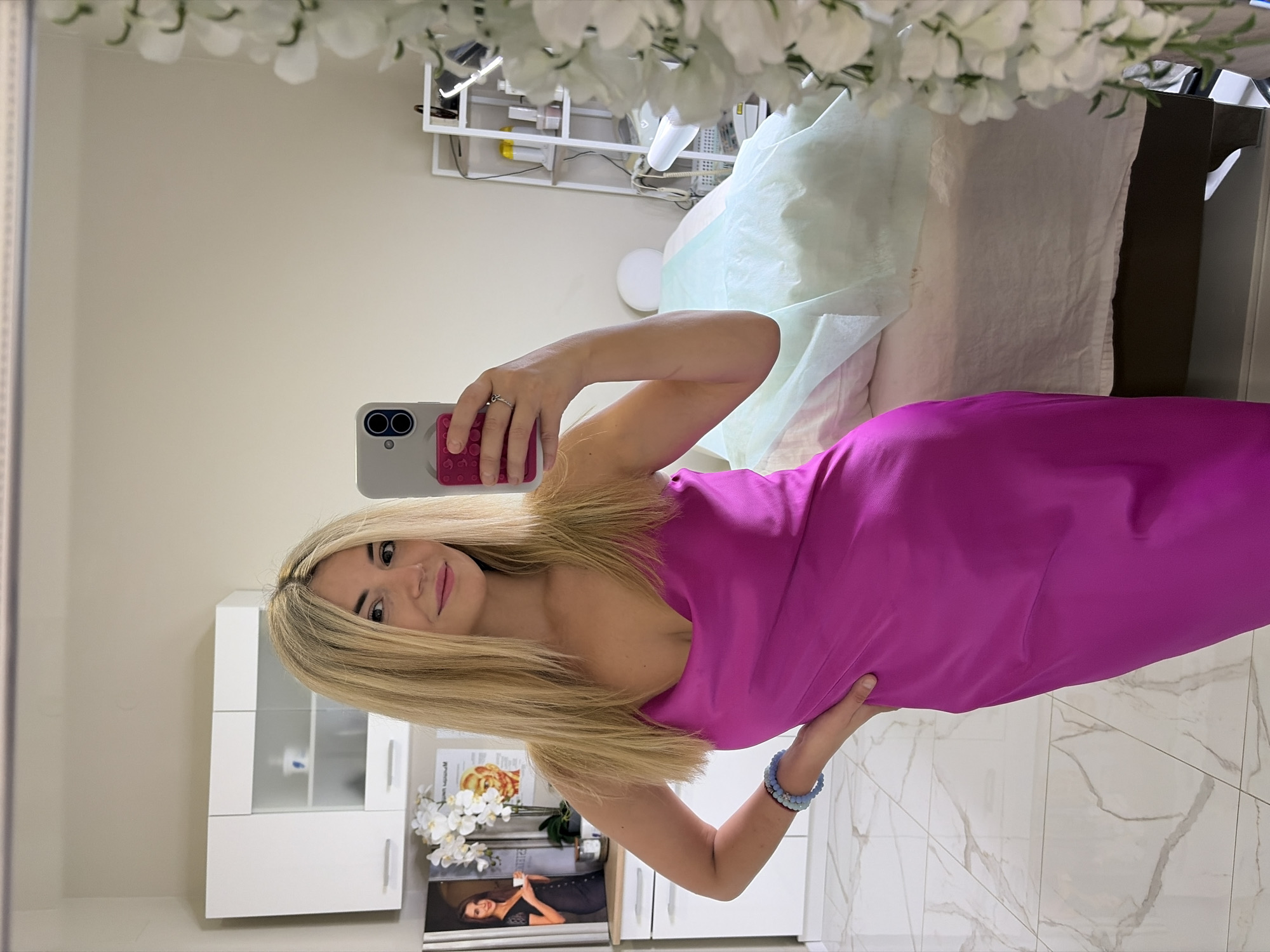 A woman with blonde hair takes a selfie in a room that appears to be a spa or treatment room. She is wearing a bright pink slip dress and has a blue beaded bracelet on her left wrist. In the background, there is a massage table covered with a light green sheet, a white shelf unit with various items, and white cabinets on the wall. There are also decorative white flowers to the right of the frame. The floor is tiled with a marble-like pattern. The mirror reflection shows the woman is holding a smartphone with a pink phone case. The overall impression is of a clean, well-lit, professional space. The context of Budva, Montenegro, suggests this might be a beauty salon or wellness center in that location.