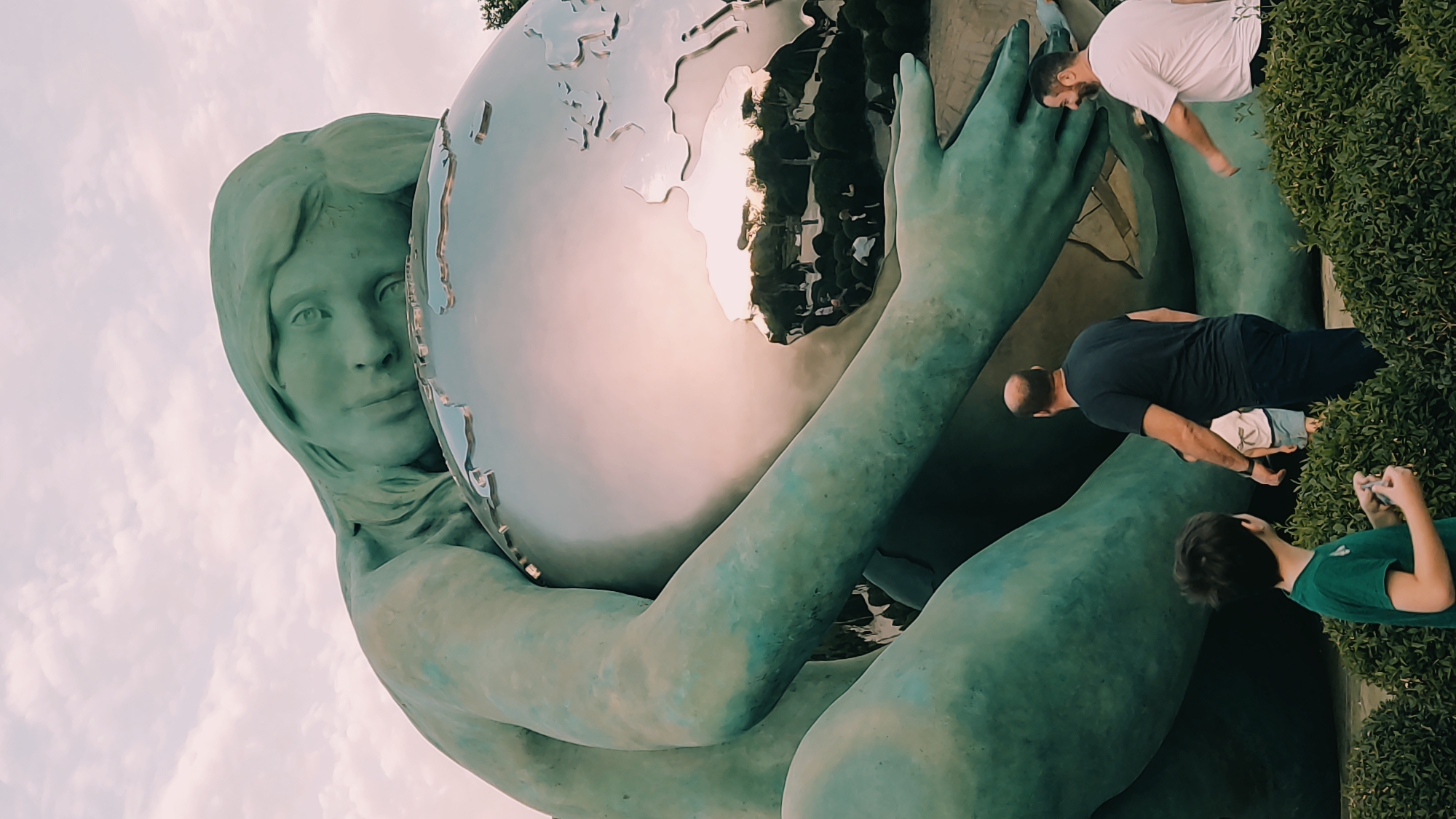 The image shows a large statue of a woman embracing the Earth. The statue is green and appears to be made of bronze or a similar material. The Earth is depicted as a reflective sphere with continents outlined on its surface. In the foreground, there are three people. On the left, a young boy in a green t-shirt is looking away from the camera. In the middle, a bald man in a black t-shirt and dark pants is walking away from the camera. On the right, a man in a white t-shirt is walking towards the camera. The background consists of a cloudy sky and some trees and bushes. This statue is located in Krasnodar, Russia, in a park called "The Gift of the Sower." The statue is a symbol of fertility and the bounty of nature.