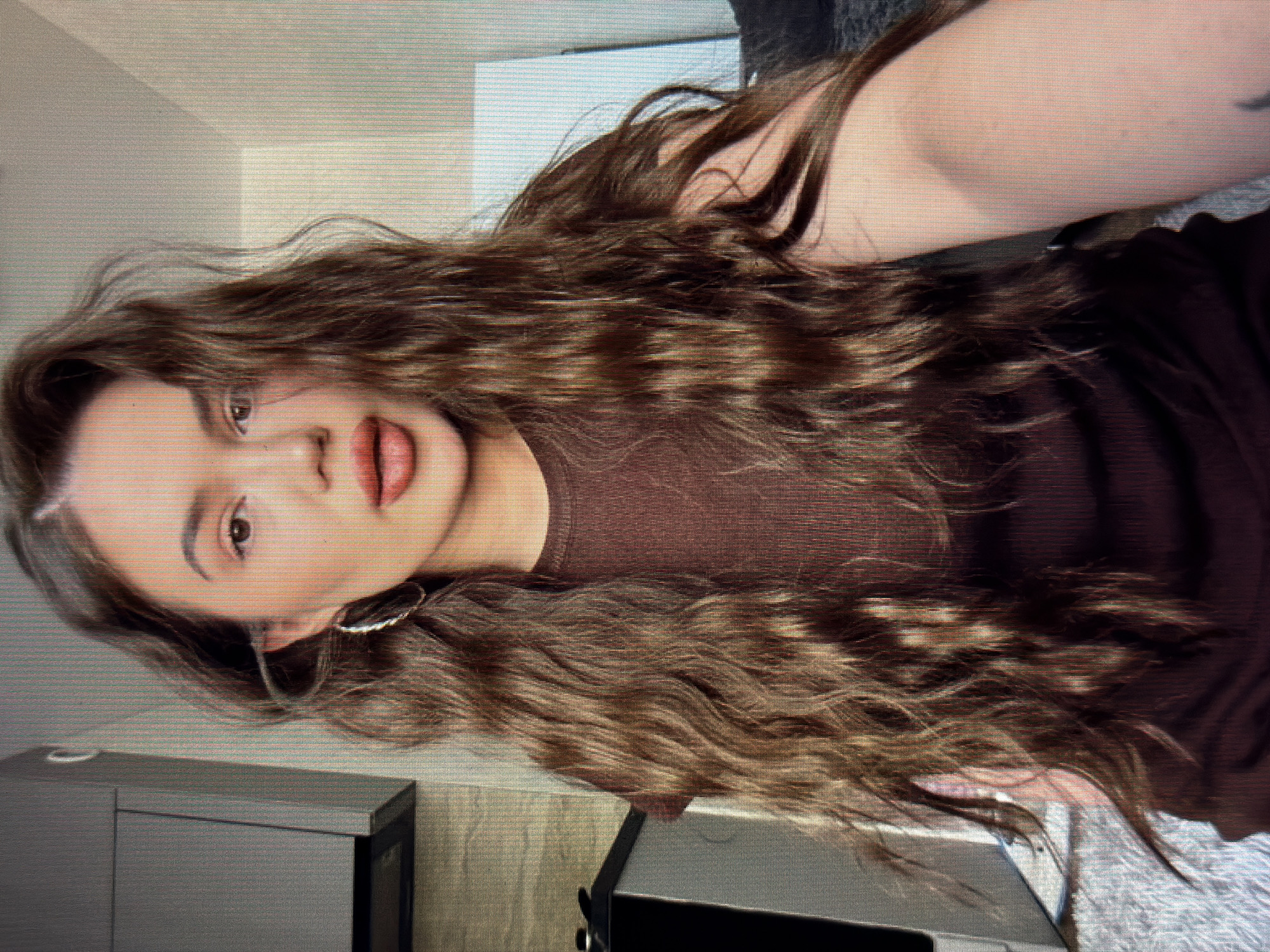 The image shows a woman with long, wavy brown hair, wearing a brown shirt and hoop earrings. She is looking towards the camera with parted lips. The background appears to be an indoor setting, possibly a kitchen or living room, with cabinets and a wall visible. There is a rug on the floor. No specific details about the location in the city of Sirius, Russia, are discernible from the image itself.