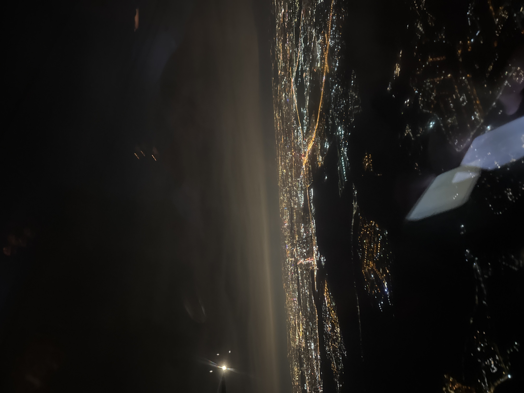 The image shows a nighttime aerial view of a city, possibly Khimki, Russia. The city is illuminated with bright lights that form linear patterns likely representing roads and densely packed areas indicating residential and commercial zones. The left side of the image is dark with some faint lights, possibly indicating a less urbanized area or a body of water. A portion of the airplane wing is visible in the upper right corner. Overall, the image is a low-angle perspective shot of a brightly lit cityscape at night.