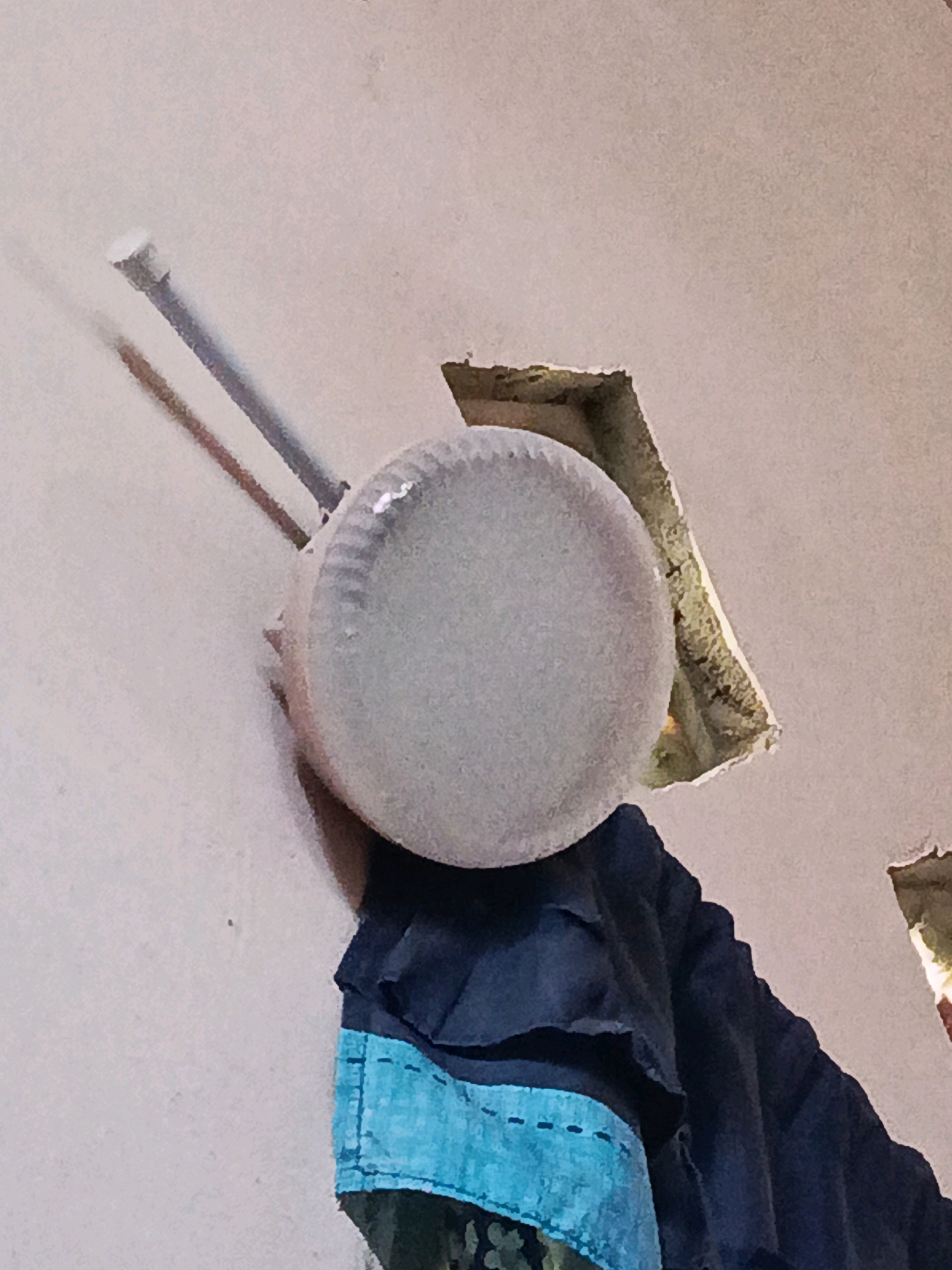 The image depicts an indoor scene with a light-colored, textured wall. Two metal pipes extend diagonally across the upper left portion of the frame, one appearing to have a white cap at its end. In the center of the image, a round, translucent object, possibly a light fixture or a bowl, hangs from or is attached to the wall. Below this object, dark blue fabric, possibly clothing, is draped. A strip of bright blue fabric with stitched details is visible on the lower part of the dark blue fabric. A rectangular opening in the wall, revealing a darker interior with some textured material, is present in the upper right and lower right of the image. The scene appears to be within a residential or utilitarian space in Megang Sakti I, Indonesia, given the context provided.