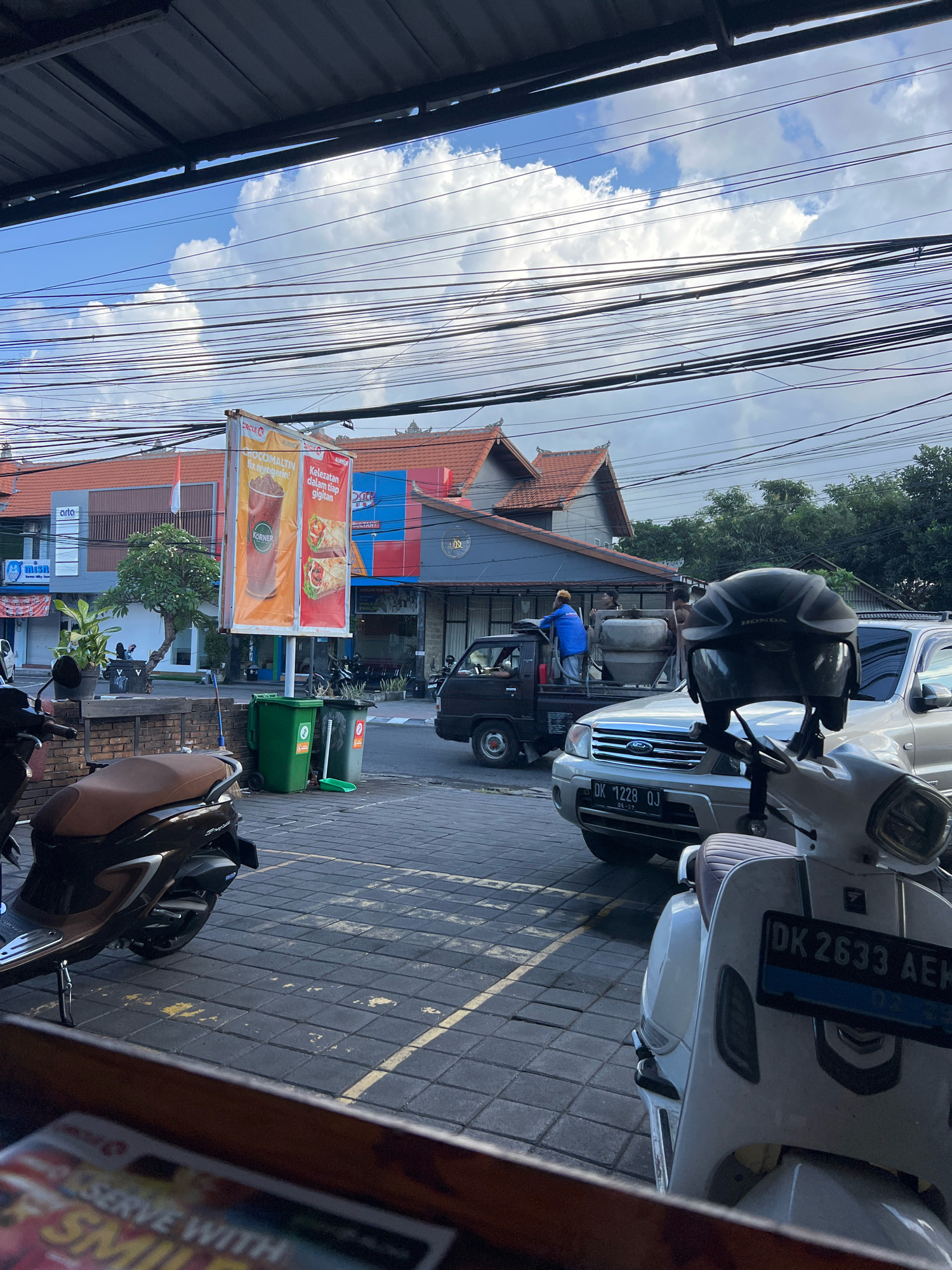 In Denpasar, Indonesia, a street scene unfolds under a bright, partly cloudy sky, with a dense network of overhead electrical wires crisscrossing the frame. The foreground is dominated by the front section of a white scooter, its license plate reading "DK 2633 AEK". To its left, a brown scooter is parked. Behind these, a silver SUV with license plate "DK 1228 QJ" is positioned, and to its left, a dark truck carries a concrete mixer. A man in a blue jacket is visible on the truck, near the mixer. Further back, shops with orange and blue signage line the street, featuring advertisements for food items. Two green trash bins and a broom are situated near the sidewalk. The street is paved with interlocking gray bricks, and a yellow line is faintly visible on the ground. Trees and buildings with red-tiled roofs contribute to the urban landscape.