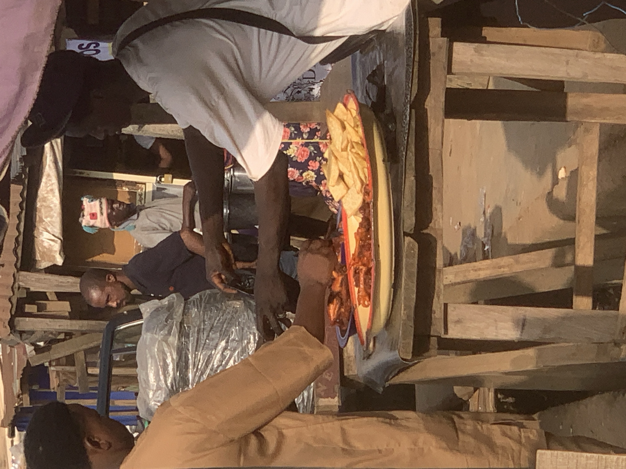 In the city of Shagamu, Nigeria, a street vendor is serving food from a stall. The vendor, wearing a white shirt and a dark cap, is standing behind a wooden table laden with food. In front of him are two platters: one filled with fried plantains and another with what appears to be fried chicken or fish. To his left, a customer in a tan shirt is reaching out to receive food. The background shows a busy marketplace with corrugated metal roofing, wooden structures, and other people. The scene is brightly lit by sunlight, casting shadows on the ground. The overall atmosphere suggests a lively commercial setting.