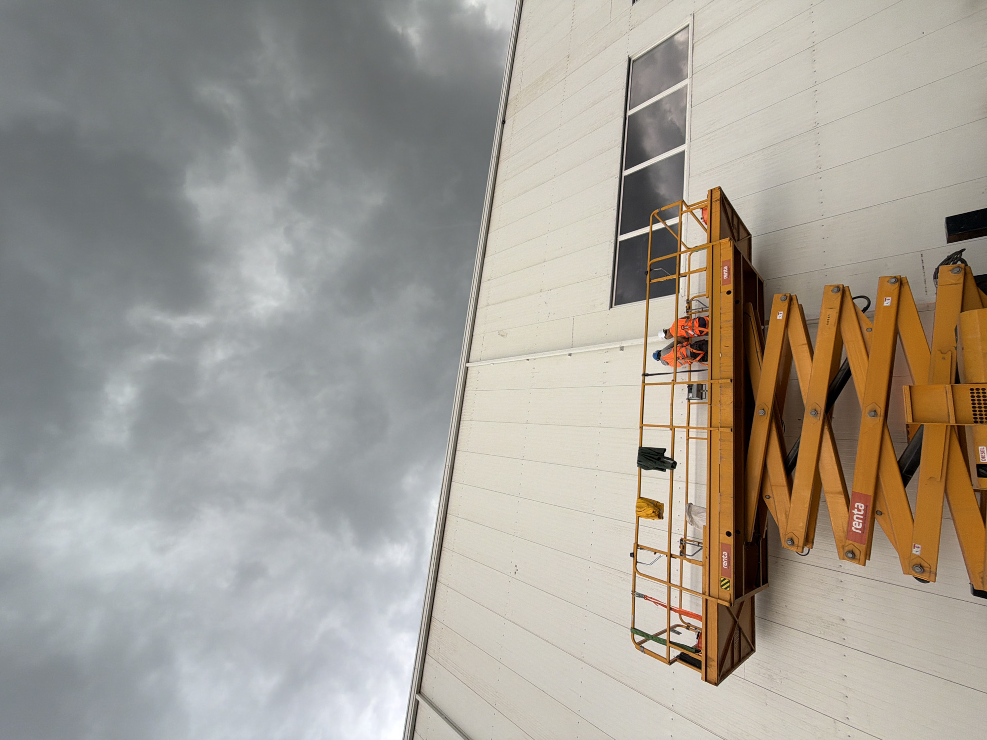 The image depicts a construction scene in Stróża, Poland, under a cloudy, overcast sky. A large, white, panel-clad building dominates the background, with a row of large, dark-paned windows positioned high on its facade. In the foreground, an orange scissor lift is raised, with its platform positioned against the building's wall. Two construction workers, wearing orange safety vests and helmets, are on the platform. One worker appears to be looking at the building wall, while the other is engaged in some activity. Several items, including a yellow garment and a dark garment, are hanging from the railing of the platform. The scissor lift has the brand name "renta" visible on its side. The overall impression is one of ongoing construction or maintenance work in potentially inclement weather.