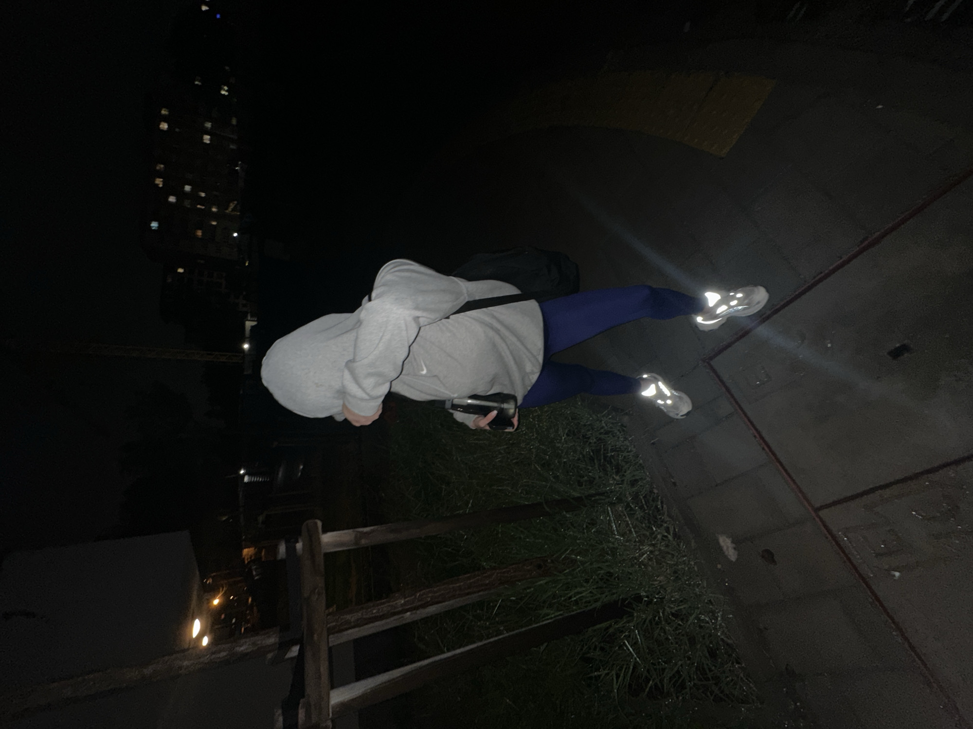 A person wearing a gray hoodie, blue pants, and reflective running shoes is standing on a sidewalk next to a patch of grass and a wooden fence. The person is holding a phone in their hands. In the background, there is a tall building with lights on in the windows. The location is Sofia, Bulgaria, as indicated by the architecture of the building in the background. The scene appears to be at night.