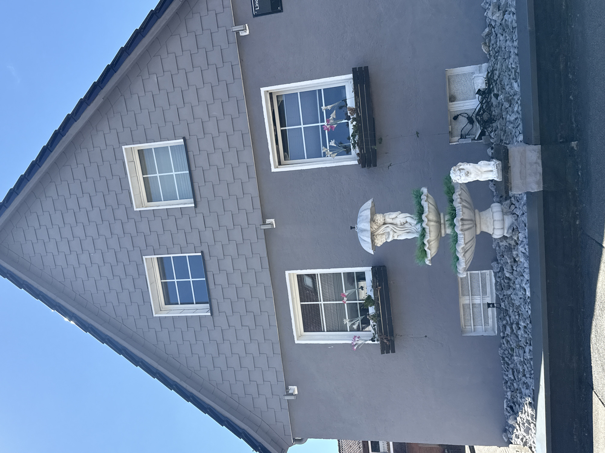The image shows the exterior of a two-story gray house located at **Steinstrasse 12, 37441 Bad Sachsa, Germany**. The house has a gray tiled roof and several windows. On the lower level, there is a decorative fountain featuring cherubs and a lion statue, surrounded by gray gravel. Two windows on the lower level are visible, one with a flower box containing pink and white flowers. Above these, two more windows are visible on the upper level, with one also having a flower box with flowers. The sky is clear and blue. The overall impression is a well-maintained residential property with decorative elements.