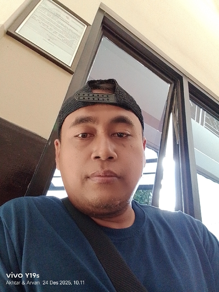 A man with a dark complexion wearing a blue shirt and a black cap looks directly at the camera. He is positioned indoors, near a window with a view of the outside. A framed document hangs on the wall behind him. The lower left corner of the image includes a phone watermark indicating "vivo Y19s" and "Akhtar & Arvan 24 Des 2025, 10.11".