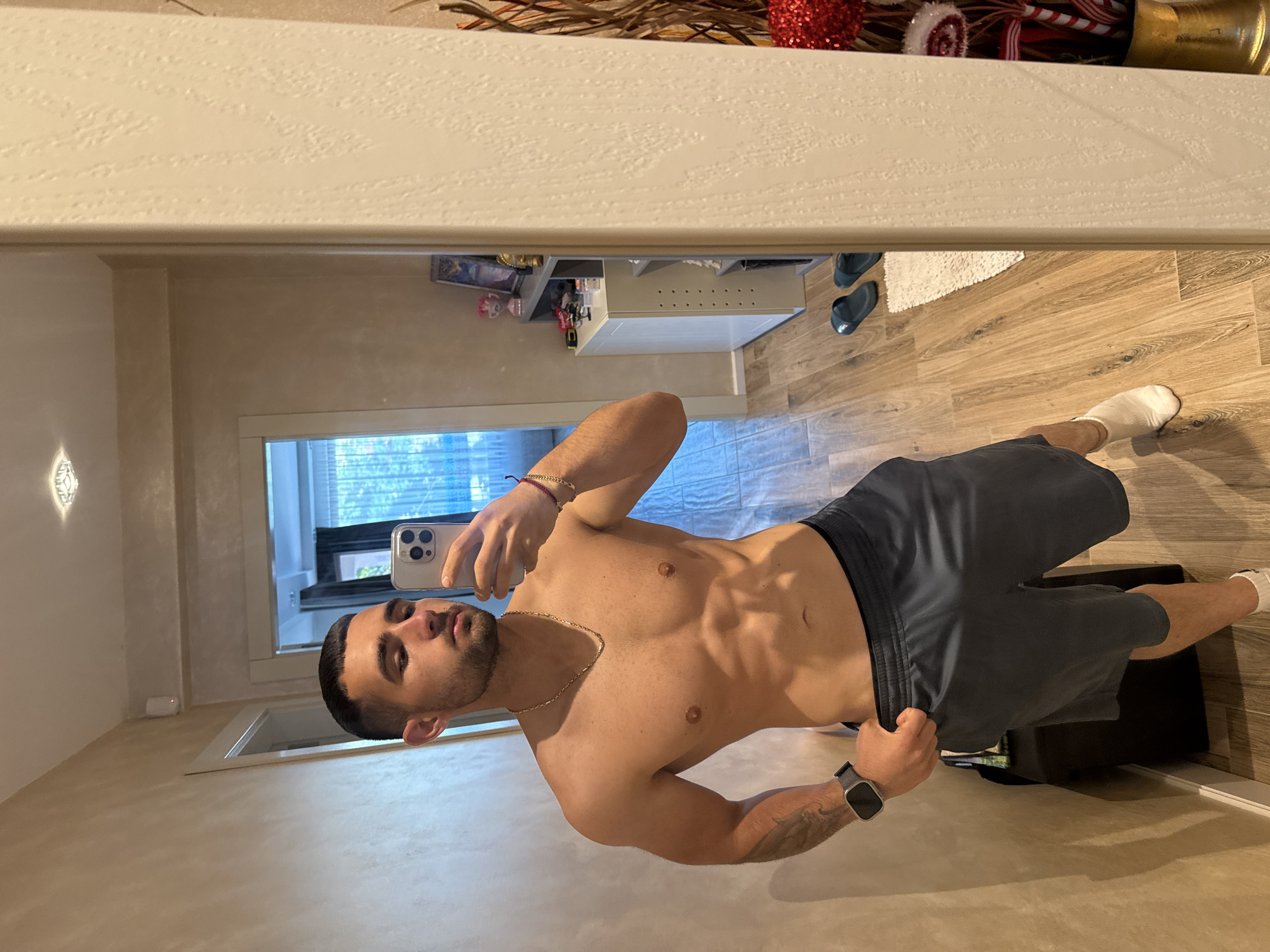 A shirtless man is taking a mirror selfie. He is wearing shorts, a watch, and a necklace. He has a short haircut and a beard. The background includes a door, a dresser, and wood flooring. The location is not discernable.