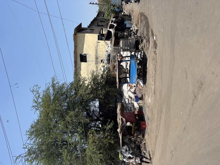 The scene depicts an outdoor urban environment, specifically a street corner or open area within Ulhasnagar, India, under a clear blue sky. Overhead utility wires traverse the upper portion of the frame. The ground consists of a dusty, unpaved or poorly maintained road surface with visible tire tracks and compacted dirt.
On the left side, a large, leafy green tree dominates the background. To the right of the tree, a two-story building with pale yellow or cream-colored exterior walls is visible. The upper story features a single window with a dark frame and a metal security grate. Adjacent to this building on the right, and extending further right, are a series of darker, more rudimentary structures, some with visible corrugated iron roofing, which appear to be extensions or smaller shanties.
In the midground, two distinct vendor carts are present. The cart on the left is painted red, has a flat, dark canopy, and is positioned near two red plastic crates on the ground. The cart on the right is painted blue, also features a dark canopy, and has a stack of three black cylindrical water tanks or containers adjacent to it.
Significant accumulations of material are present throughout the scene. Large piles of white or off-white sacks, likely containing goods or recyclables, are stacked against the buildings and around the base of the large tree. Interspersed within these piles and scattered on the ground are various items, including dark tarps, scrap materials, plastic debris, and general refuse. A blue plastic crate is observed near the blue cart.
One individual, dressed in a light-colored shirt and dark pants, is visible on the left side of the road, near the red cart. This person is captured in a dynamic pose, facing right, with their right arm raised and left arm lowered, suggesting movement or an active task. A second individual is partially visible on the far right edge of the image, seated on a dark-colored motorcycle and moving away from the viewer. The scene is brightly illuminated by natural daylight.