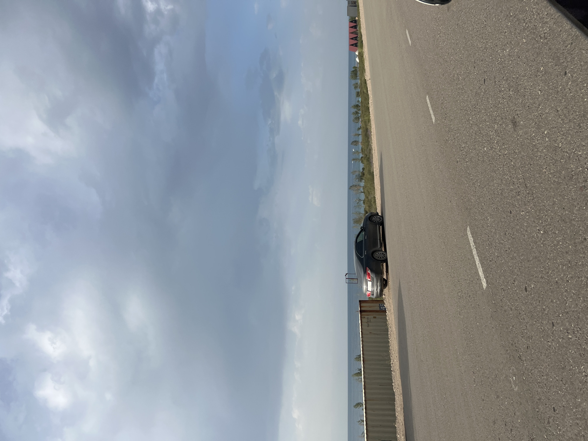 An overcast sky hangs above a paved road in Kaji-Say, Kyrgyzstan. A dark sedan is parked on the shoulder of the road, facing away from the camera, near a large, corrugated metal shipping container. Beyond the container, the blue waters of Issyk-Kul lake stretch to the horizon, where a line of trees and small red-roofed structures are visible. The road has white dashed lines marking the lanes.