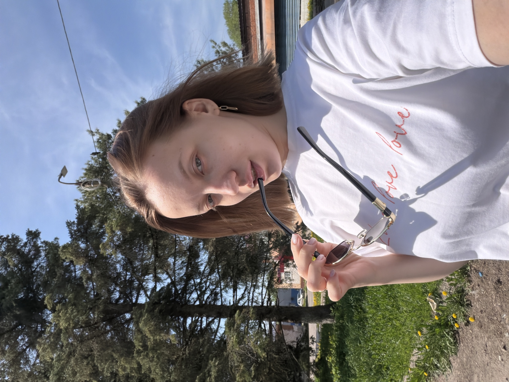 A young woman with light brown hair and fair skin stands outdoors on a sunny day. She wears a white t-shirt with the words "we love" written in red script. She is holding a pair of sunglasses with a black frame and gold accents, partially held in her mouth. The background includes a tall evergreen tree, green grass dotted with yellow dandelions, a building with a red brick facade, and a blue sky with scattered clouds. The location is in Reshetnikovo, Russia.