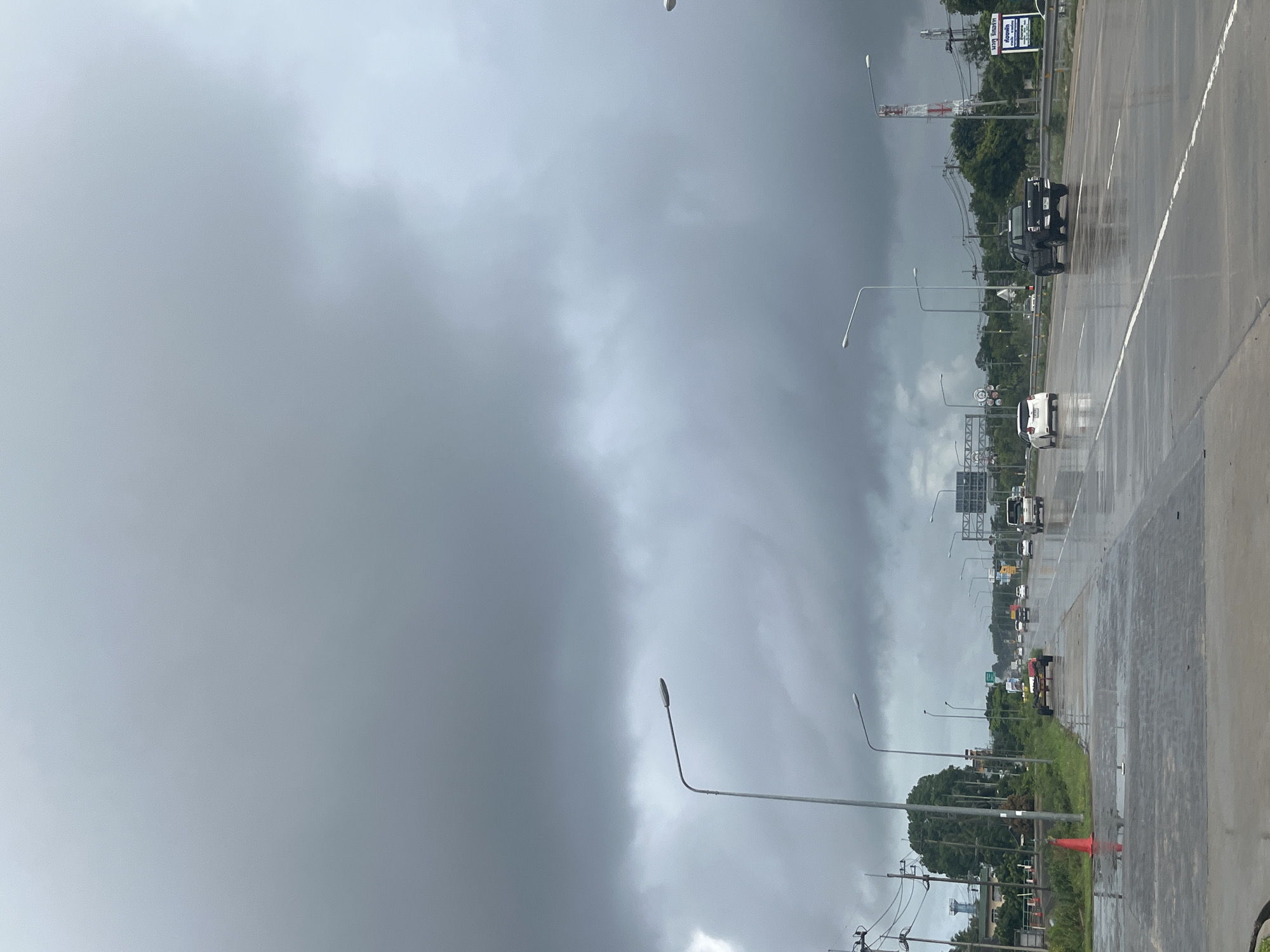 The image depicts a wet, overcast day on a highway in บ้านหนองน้ำเต้าลอย, Thailand. The sky is filled with dark, ominous clouds. The road surface is wet, reflecting the sky and vehicles. There are vehicles on the road, including a white car, a black pickup truck, and other unidentified vehicles further down the road. There are streetlights along the side of the road. Beyond the road, there is green vegetation and utility poles. There are also road signs visible.