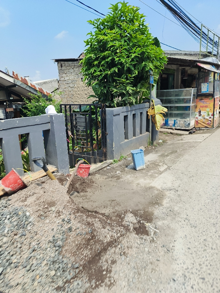 The image shows a scene in Padalarang, Indonesia, located at latitude -6.851 and longitude 107.475. A pile of gravel sits in the foreground, with a shovel resting on top of it. A blue plastic bucket sits on the ground to the right of the gravel. There is a red bucket to the left of the gravel pile, and a partially obscured person is visible in the background. A small tree grows in the center of the scene. There is a fence to the left of the gravel, and a small building to the right. The image was taken in daylight.