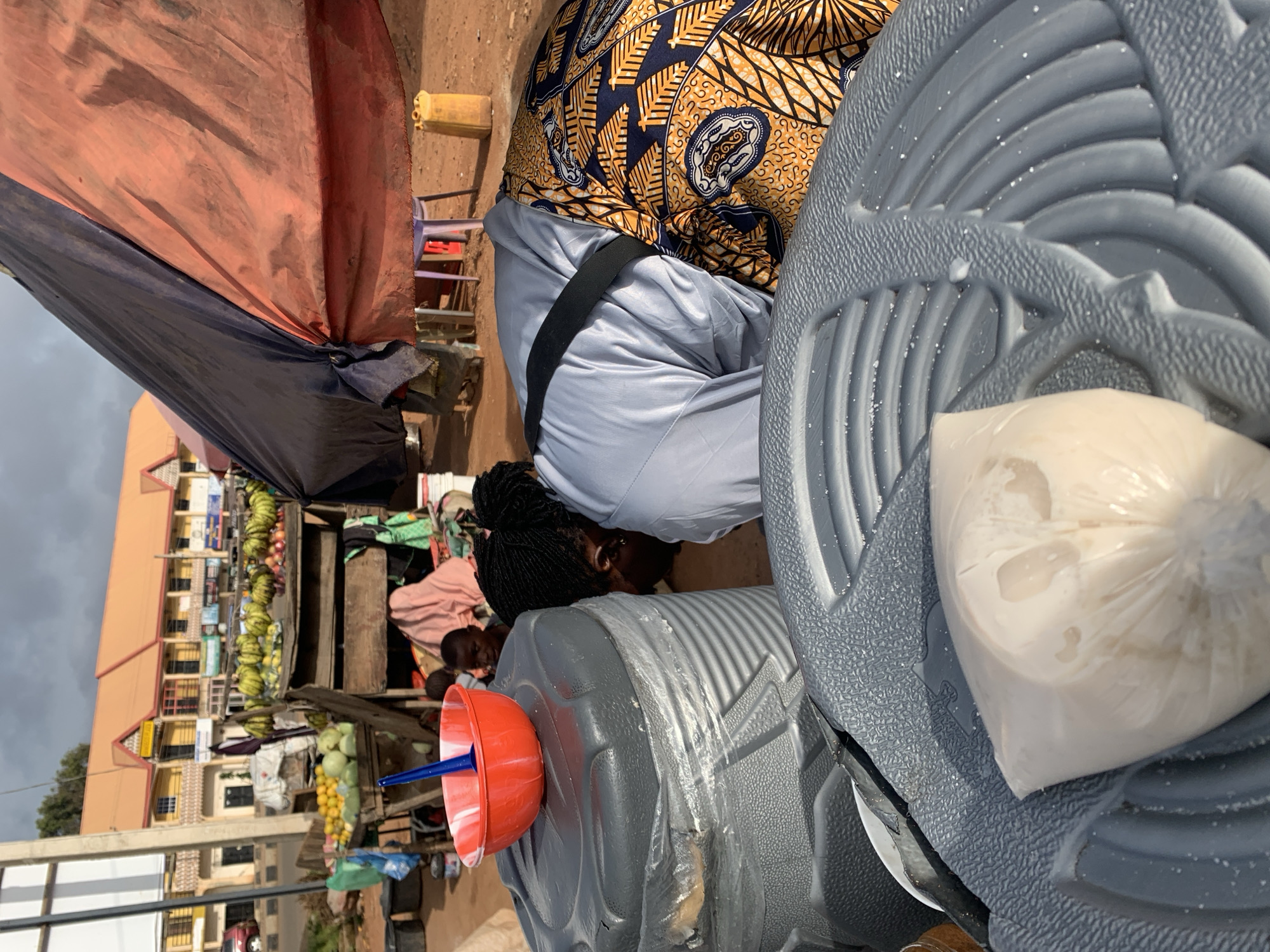 The image depicts an outdoor market scene in Gura Top, Nigeria, under a cloudy sky. In the foreground, a person wearing a light blue shirt and a vibrantly patterned yellow and black wrap skirt is bent over, facing downwards, with their head obscured. A black strap is visible across their back. They are positioned near large, grey, ribbed plastic containers, one of which has a red bowl with a blue spoon resting on its lid. A plastic bag containing a pale, semi-liquid substance is placed on a grey, ribbed plastic surface in the lower part of the frame. In the midground, a stall displays an assortment of fruits, including bunches of bananas and other produce. Behind the stall, several children are visible, one of whom is partially obscured by a pink cloth. In the background, a row of buildings with orange roofs and beige facades lines a street. The ground is covered in reddish-brown dirt. A tall, white pole with a dark rectangular sign is visible on the left side of the image.