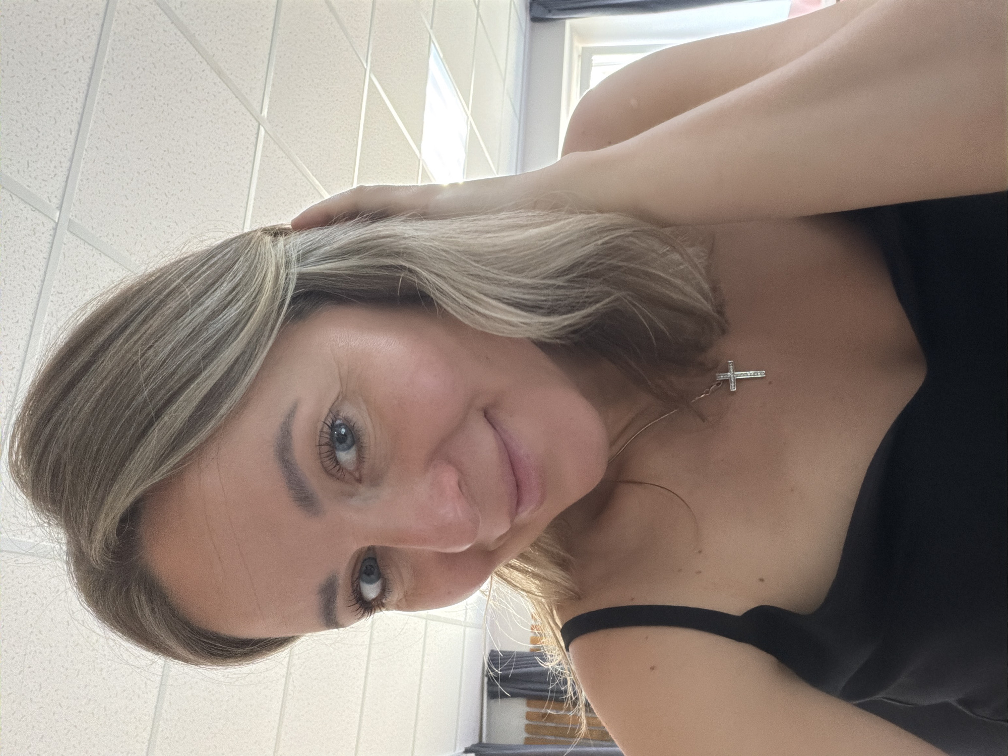 A woman with blonde hair and blue eyes is pictured in a selfie. She is wearing a black top and a silver necklace with a cross pendant. Her right hand is in her hair. The background features a white tiled ceiling and a window. The image does not provide sufficient information to determine if the location is in the city of Chaikovsky, Russia.
