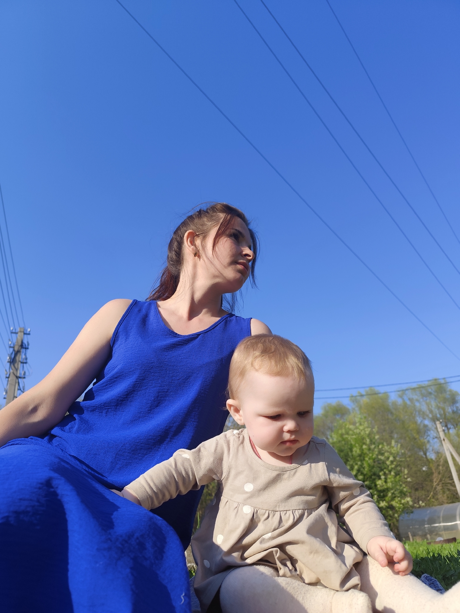In an outdoor setting in Фоки, Russia, a fair-skinned woman with brown hair pulled back in a ponytail sits beside a fair-skinned baby with blonde hair. The woman is wearing a blue sleeveless dress. The baby is wearing a light brown dress with white polka dots and light-colored tights. They are sitting on what appears to be grass. Overhead, clear blue sky is crossed by power lines supported by utility poles. In the background are trees, bushes, and a greenhouse.