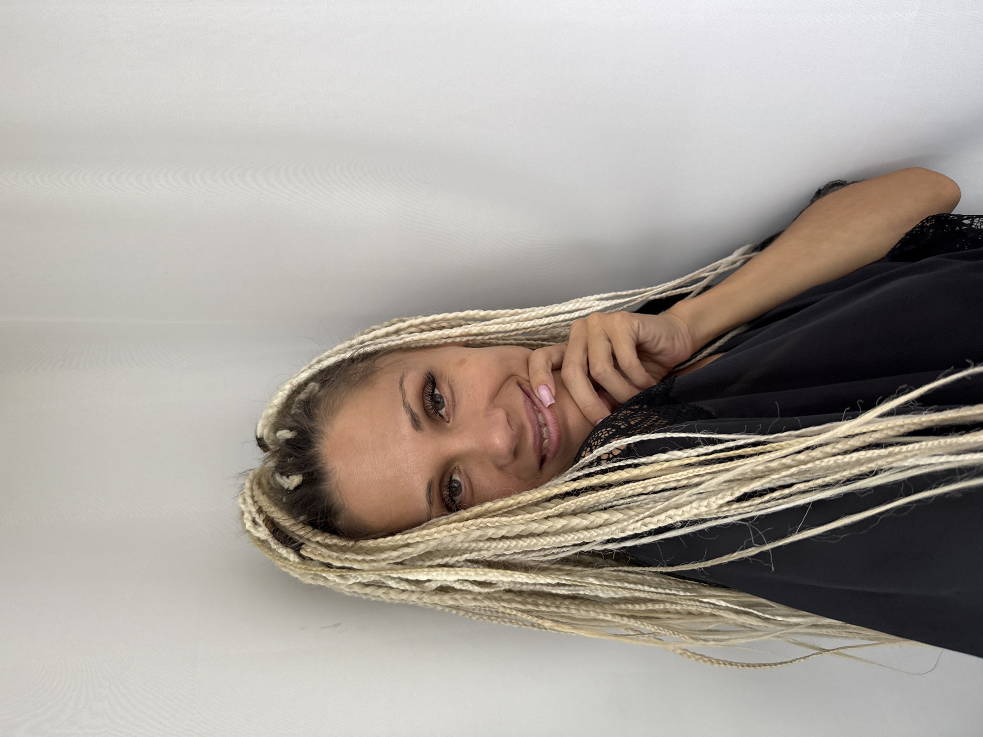 A woman with blonde braided hair is posing against a white wall. She is wearing a black garment with lace detailing. Her right hand is touching her chin, and she is smiling. The lighting is soft, and the background is a plain, textured white wall. The scene appears to be indoors, with no specific urban or natural features identifiable to pinpoint a location in Rusanovo, Russia.