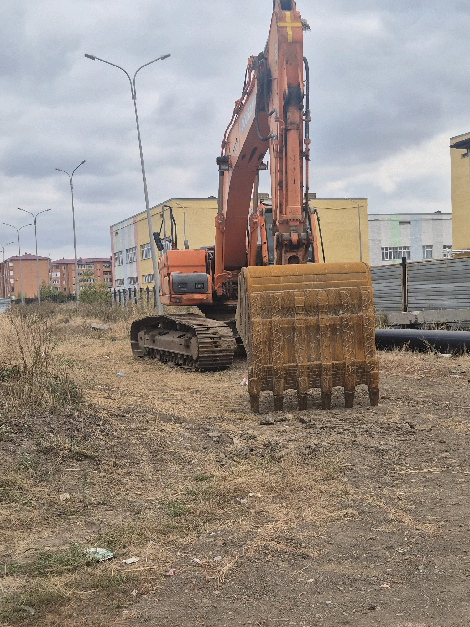 An orange excavator with a large bucket sits in a dry, grassy field. In the background, a light yellow building with colorful panels on its facade is visible, along with other apartment buildings and streetlights. The sky is overcast.