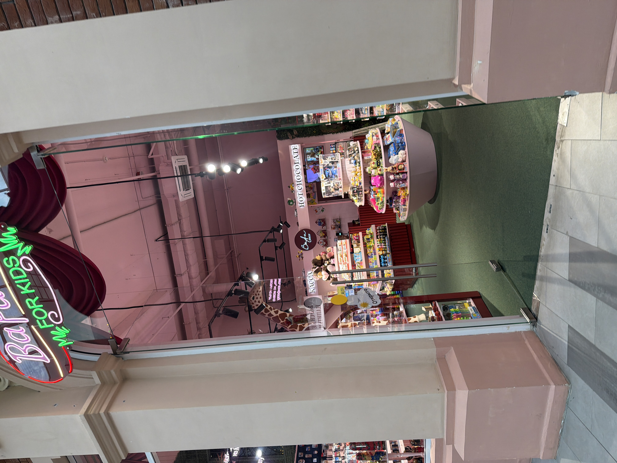 The image shows the entrance to a store with a pink and cream facade. A neon sign above the entrance reads "Bara for Kids" in pink and green lettering. Through the glass doors, the interior of the store is visible. The store appears to be a toy or candy shop, with shelves stocked with colorful merchandise. A large stuffed giraffe head is visible in the doorway. The floor inside the store is covered with green carpet, and there are display shelves filled with toys and other items. The lighting inside the store is pink. The store is located in a mall or shopping center, as evidenced by the brick wall on the right side of the image. The overall impression is one of a bright, cheerful, and inviting retail space.