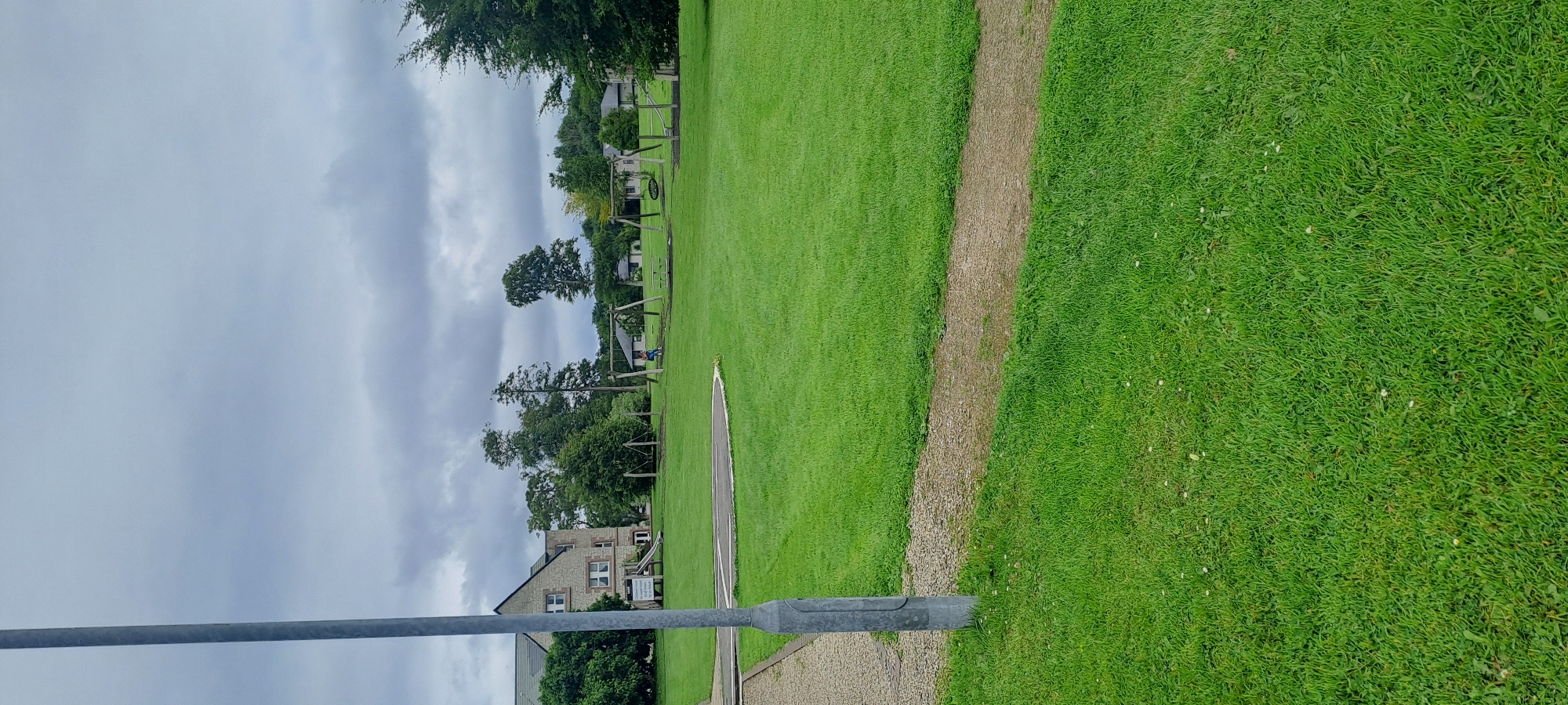 A tall, gray metal pole stands in the foreground to the left, casting a shadow on the gravel path and bright green grass. The path leads into a grassy field dotted with small white flowers. In the middle ground, a playground area features swings, a slide, and a tire swing. A person in a blue shirt is visible on one of the swings. Behind the playground, several houses and trees are scattered across the landscape. The sky above is overcast with dark, heavy clouds. The scene appears to be a public park or recreational area in Castlebar, Ireland.