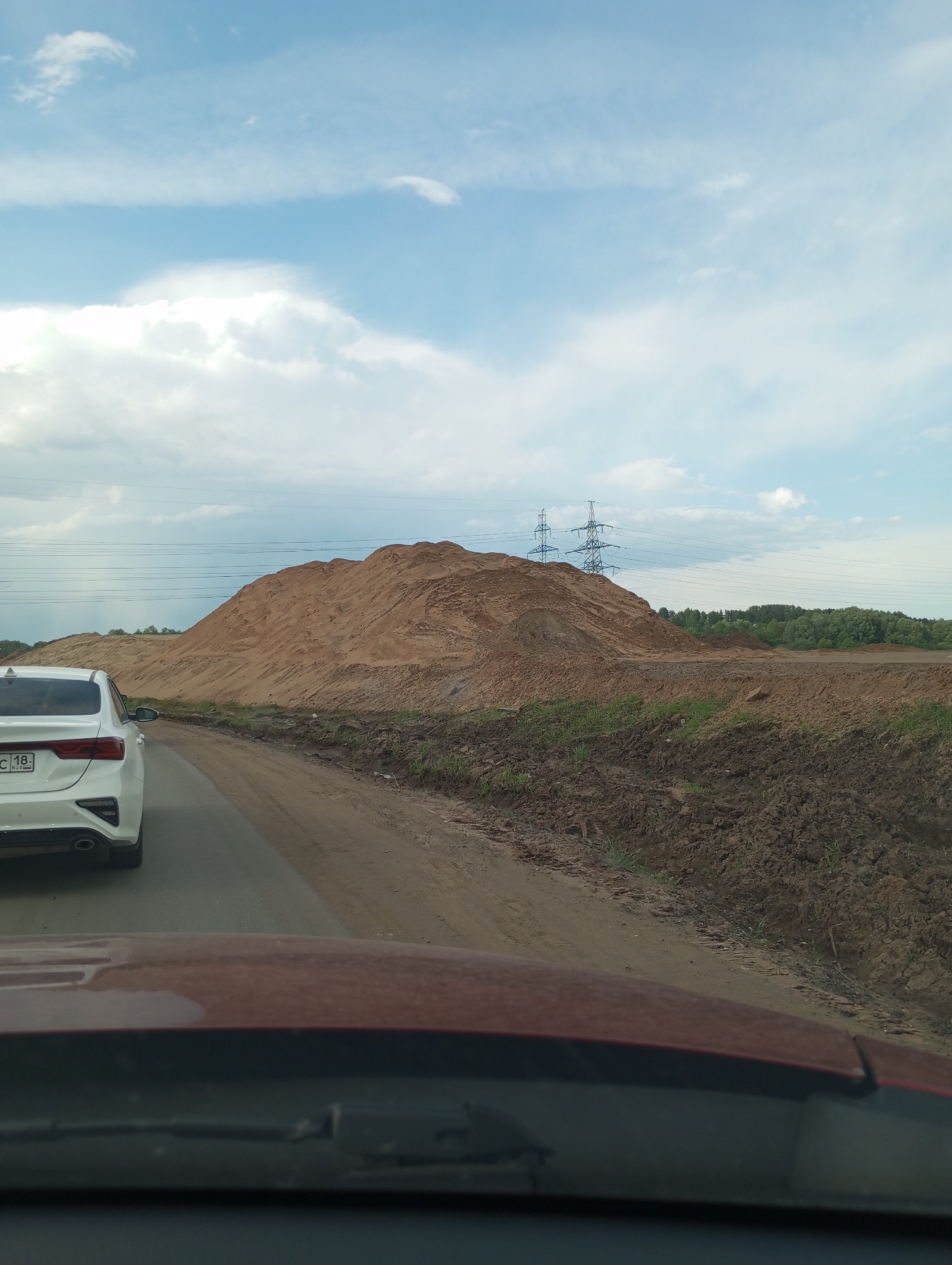 The image, taken from inside a car, shows a large mound of dirt or sand next to a road. The back of a white car is visible on the left side of the road. Power lines are present in the background, along with trees in the distance. The sky is cloudy. The vehicle's hood and windshield are visible in the foreground. The presence of Russian license plates, along with the explicit statement in the prompt, suggest the location is likely somewhere near the city of Kabanikha, Russia. Exact location is not discernible.