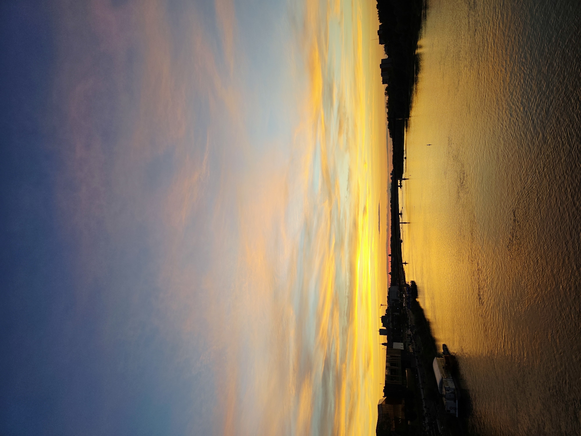 The image depicts a wide river in Warsaw, Poland, reflecting the vibrant colors of a sunset. The sky above transitions from a deep blue at the top to hues of pink, orange, and brilliant yellow closer to the horizon. Wispy clouds are visible, painted with the warm tones of the setting sun.
On the left bank of the river, a city skyline is silhouetted against the colorful sky. Buildings of various heights are visible, including what appears to be a cluster of taller structures in the center, possibly representing the business district. A dark, modern building with angular lines is prominent in the foreground on the left, and further along the embankment, a long, white boat is moored. People can be observed walking along the riverfront promenade.
The river itself is a broad expanse of water, its surface rippled by gentle waves, and it reflects the intense golden light of the sunset, creating a mirror-like effect. A bridge spans the river in the distance, with indistinct shapes of vehicles or people visible on it. On the right bank, another silhouetted city skyline is visible, with trees and buildings forming a dark line against the bright sky. The overall impression is one of tranquility and the beauty of urban nature during twilight.