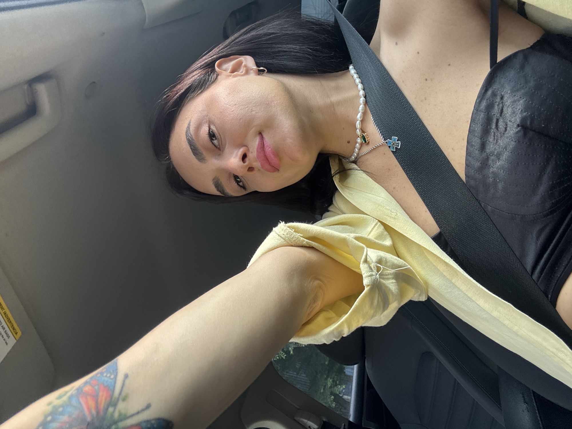 The image shows a woman taking a selfie in a car. She has a butterfly tattoo on her left arm and is wearing a light yellow shirt, a black top, and a pearl necklace with a pendant. The woman is looking at the camera with her lips slightly parted. The car interior is visible, including a black seat and a seatbelt. The background shows some greenery, suggesting the car is in a city, possibly Tbilisi, Georgia, though the location is not explicitly stated in the image. The lighting suggests it is daytime.
