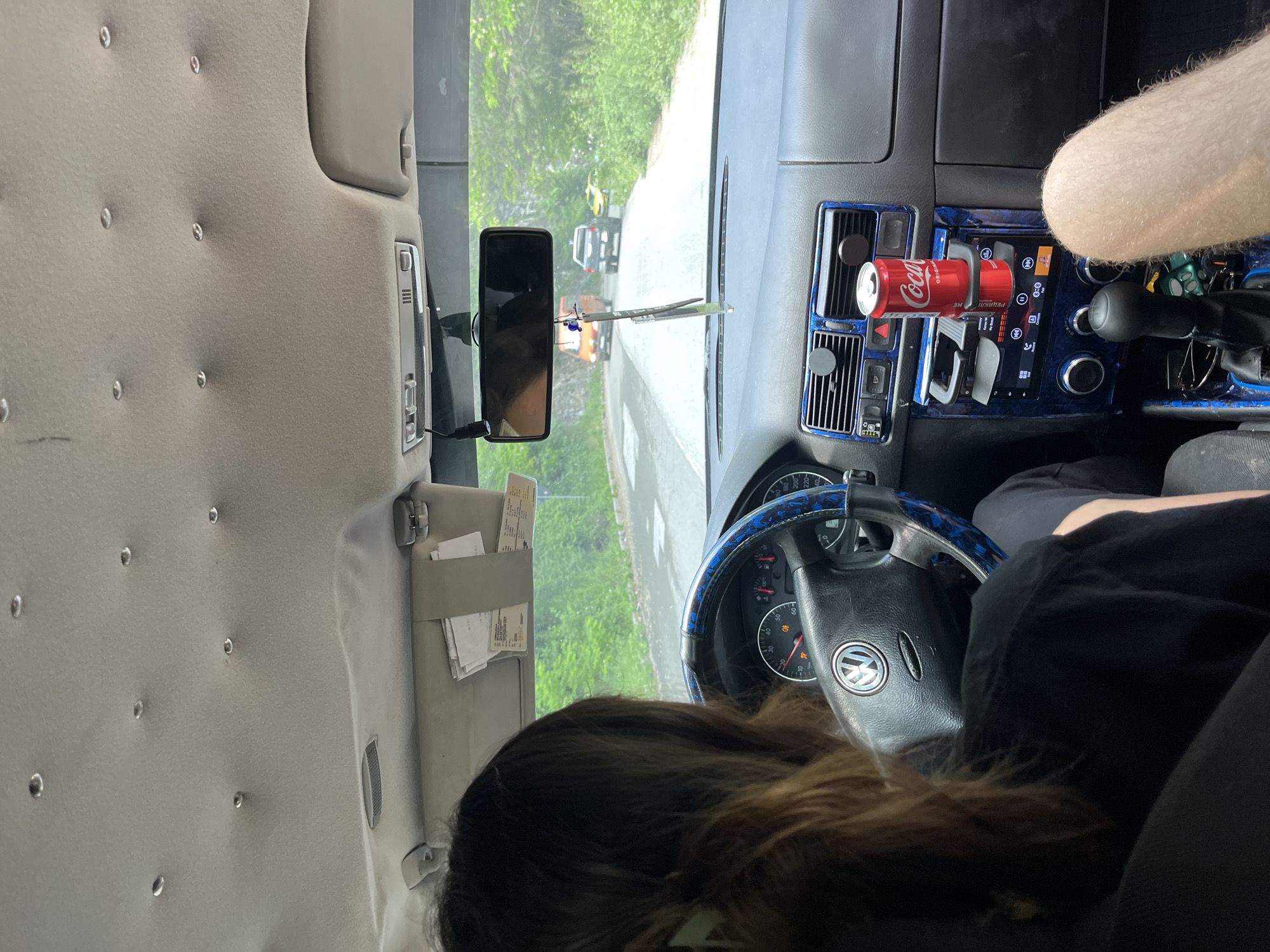 The image, taken from inside a car, shows the perspective of a passenger looking towards the driver and the road ahead. The driver, a person with long brown hair, is partially visible from the back. The car's interior is detailed, featuring a Volkswagen steering wheel with a blue patterned cover, a dashboard with air vents and a car stereo, and a can of Coca-Cola in a cup holder. A rearview mirror reflects the road, where an orange van, a dark-colored SUV, and a yellow car are visible. The exterior environment appears to be a road surrounded by lush green trees, suggesting a rural or suburban setting. The presence of the Coca-Cola can and air freshener hanging from the rearview mirror indicate the car is in use. There is no specific indication of the location being in the city of Vlahovo, Bulgaria.